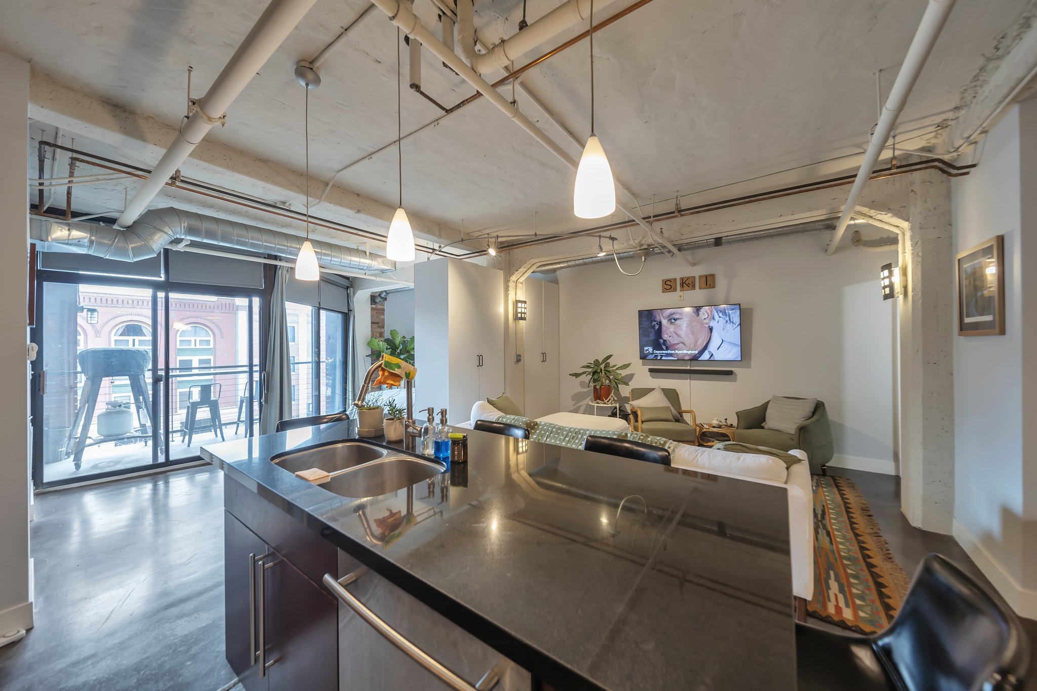 Open-concept living space featuring a kitchen island with dual sinks and dark cabinetry, exposed ceiling ductwork and piping, and dark wood-finish flooring