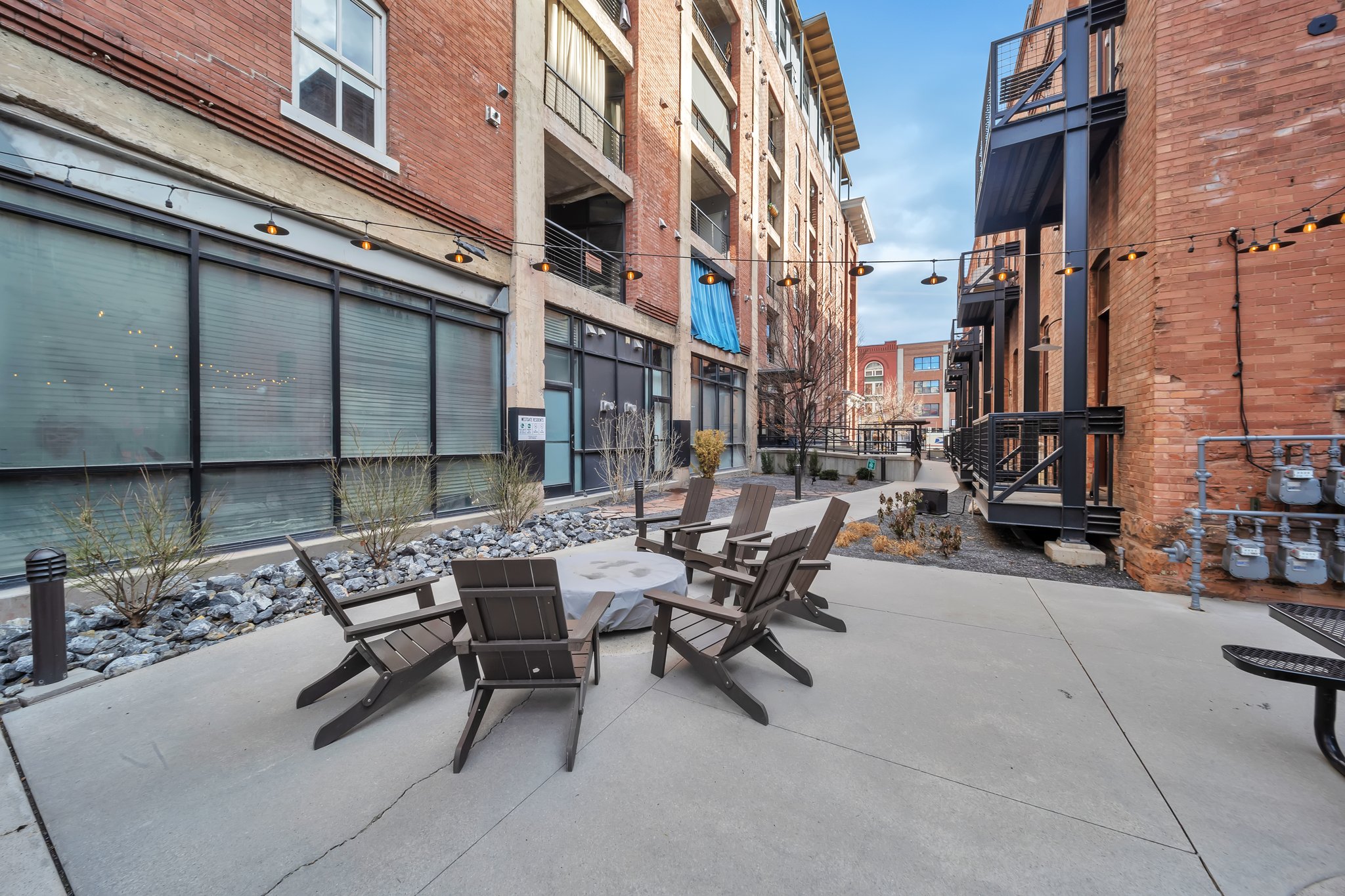 Paved courtyard featuring a central fire pit, surrounding Adirondack-style chairs, string lighting, and brick facades