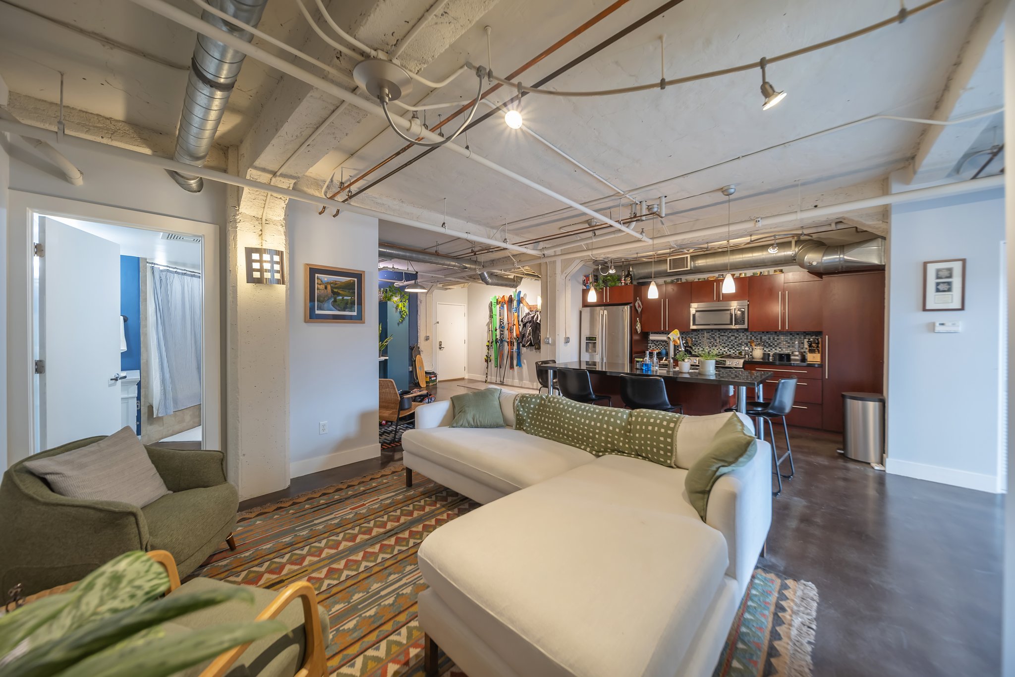 Open-concept living space featuring exposed ductwork and ceilings, concrete flooring, and a kitchen with dark wood-finish cabinetry, stainless steel appliances, and a tiled backsplash