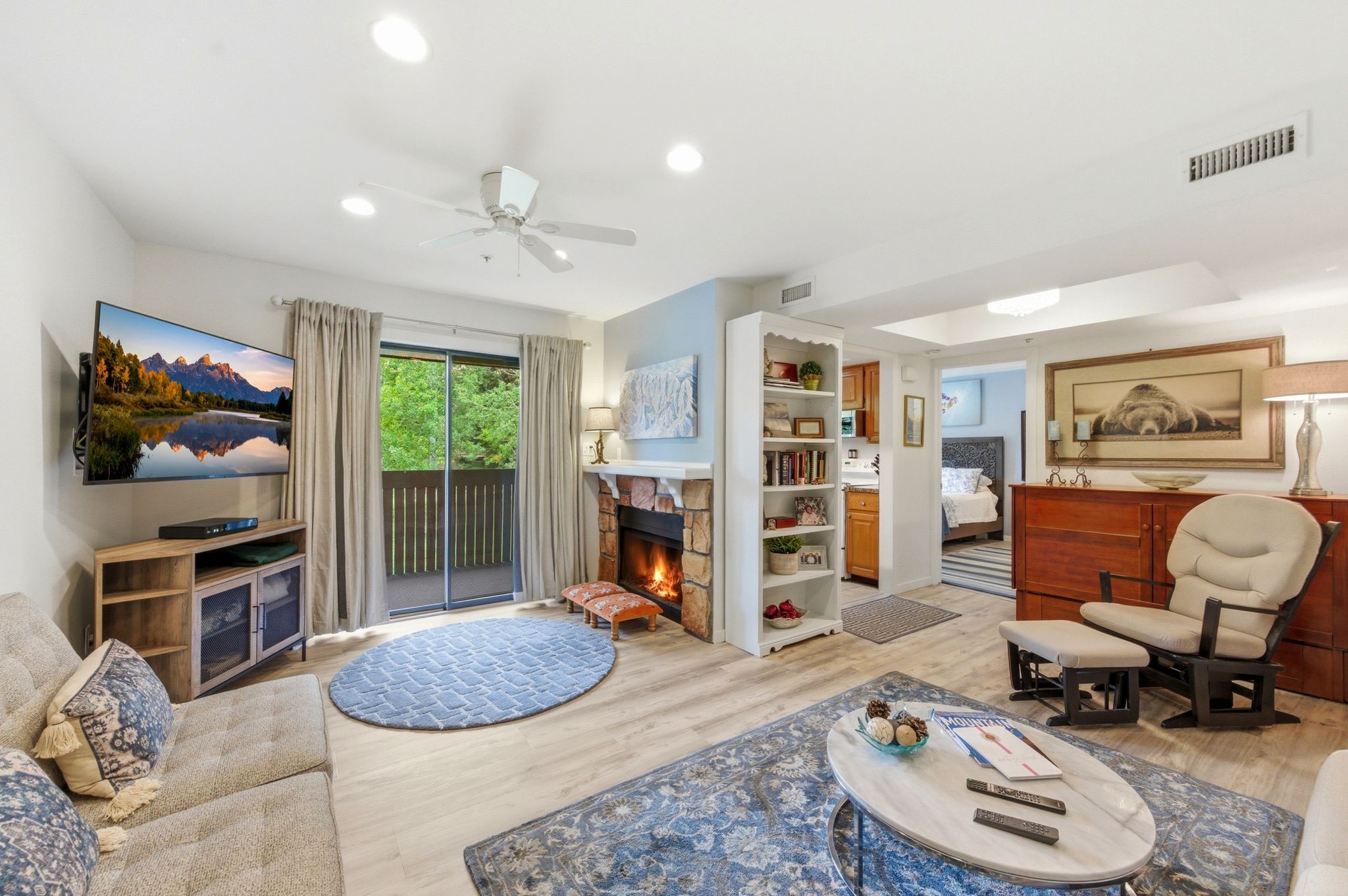 Open-concept living area featuring wood-finish flooring, a stone-faced fireplace, a ceiling fan, recessed lighting, and a sliding glass door opening to an outdoor space