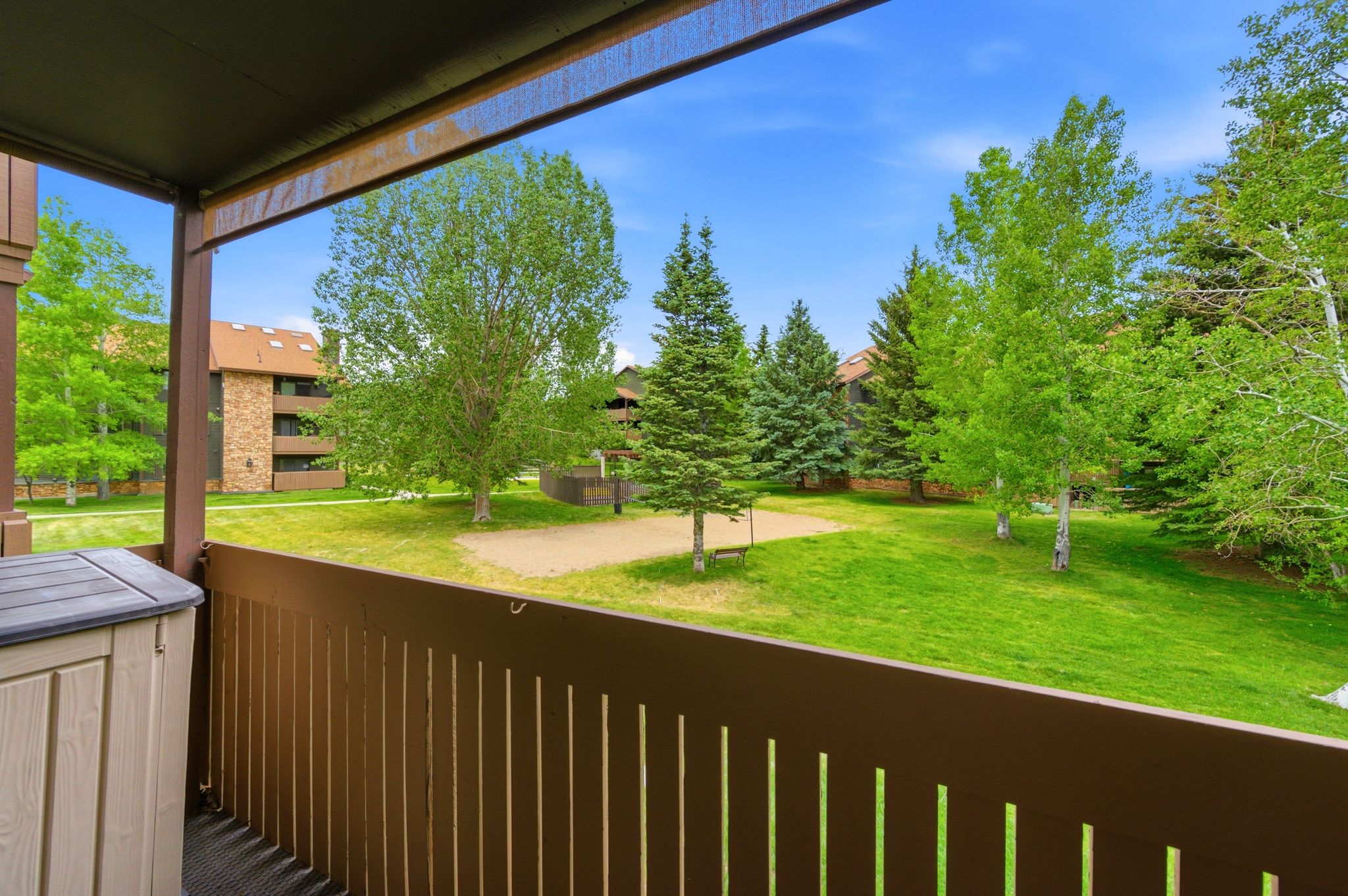 Private balcony with slatted railing, overlooking a lush green lawn, mature trees, and a sand court