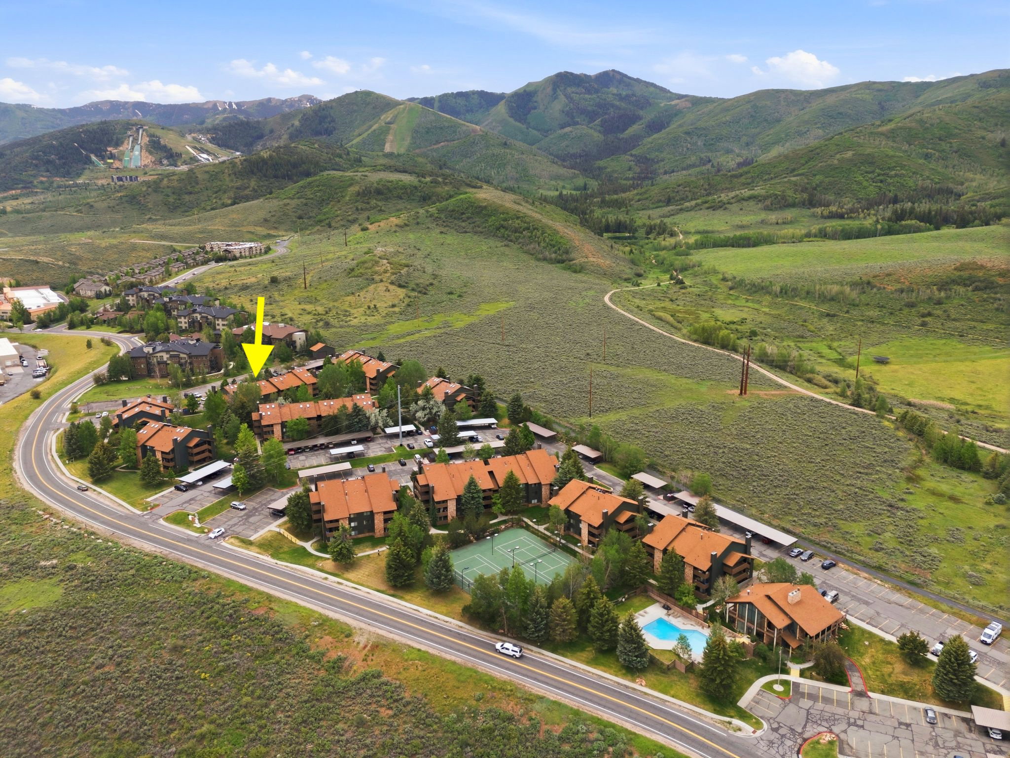 Residential community situated against a mountain backdrop, featuring multiple buildings with terracotta-style roofs, a dedicated tennis court, and an outdoor swimming pool