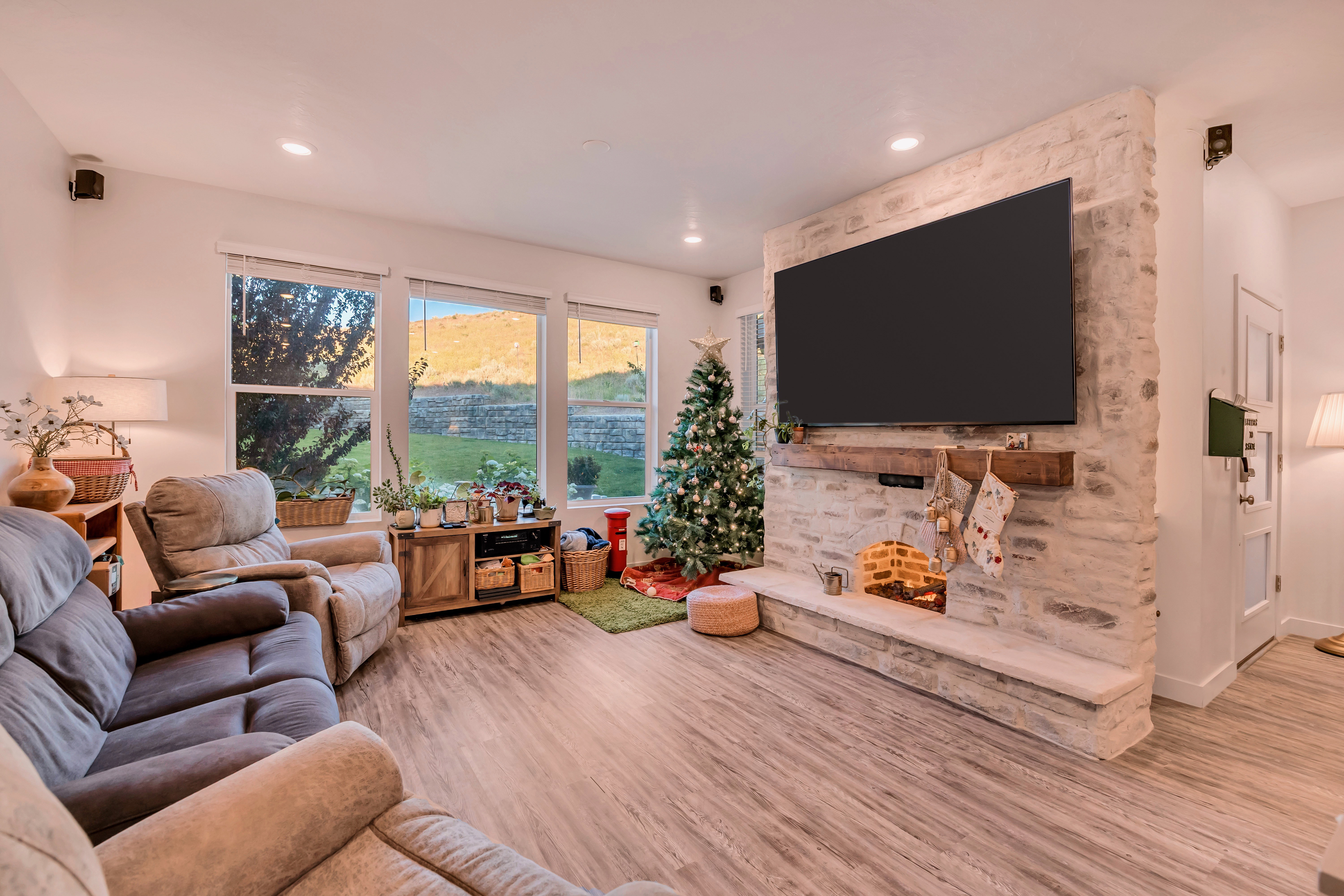 Spacious living area featuring expansive windows, wood-finish flooring, a stone accent wall, and a rustic wood mantel
