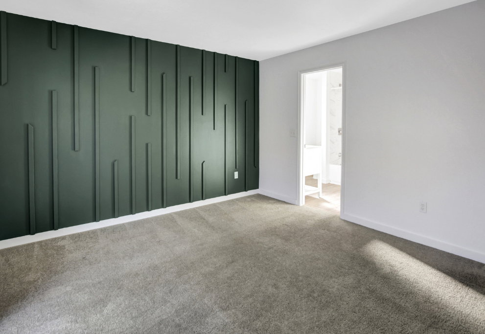 Accent wall featuring vertical paneling, light gray carpeting, white baseboards, and a doorway leading to a bright bathroom with a white vanity