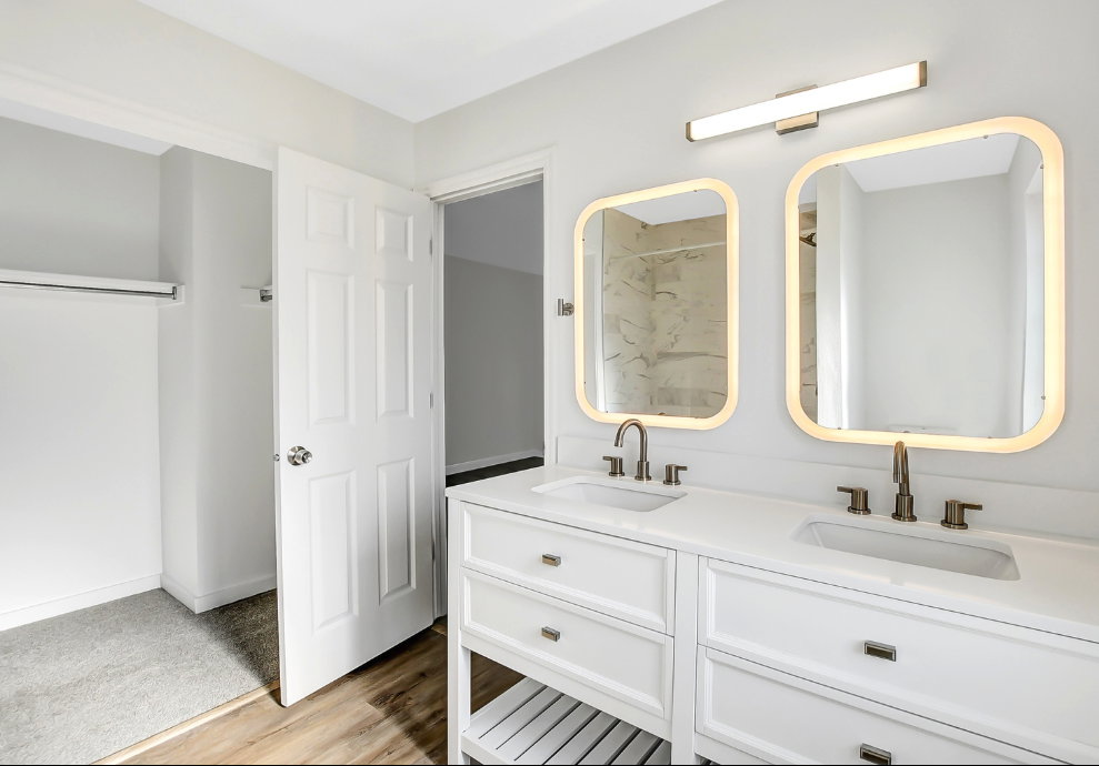 Dual vanity with white cabinetry and integrated sinks, illuminated by backlit mirrors and an overhead light bar