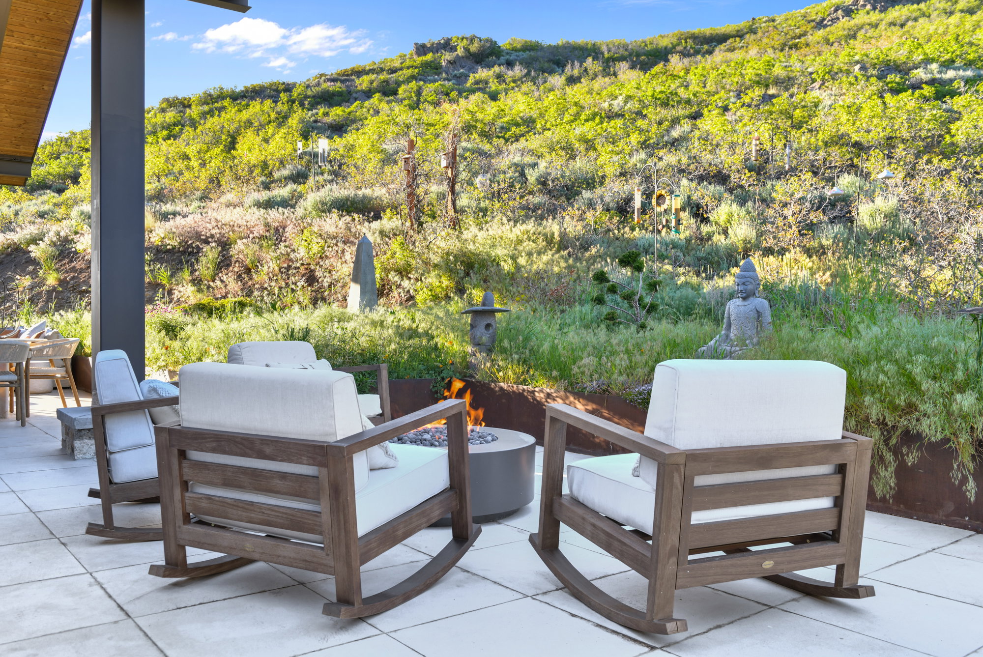 Expansive outdoor patio featuring light-toned paver flooring, a built-in fire pit, and a backdrop of natural landscaping