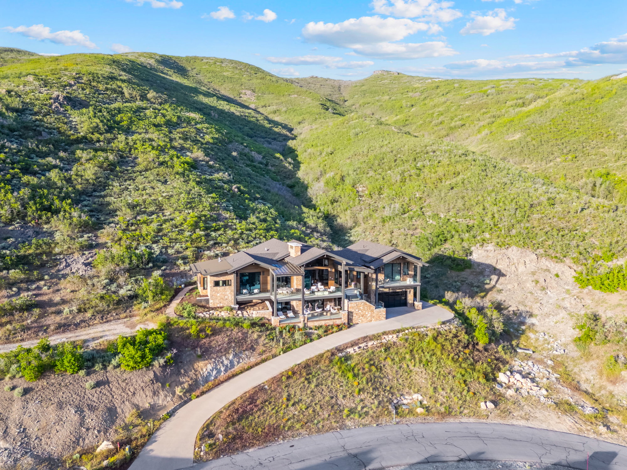 Hillside residence featuring stone and wood-clad exterior, multiple levels of balconies, a paved driveway, a multi-car garage, and a sloped terrain backdrop