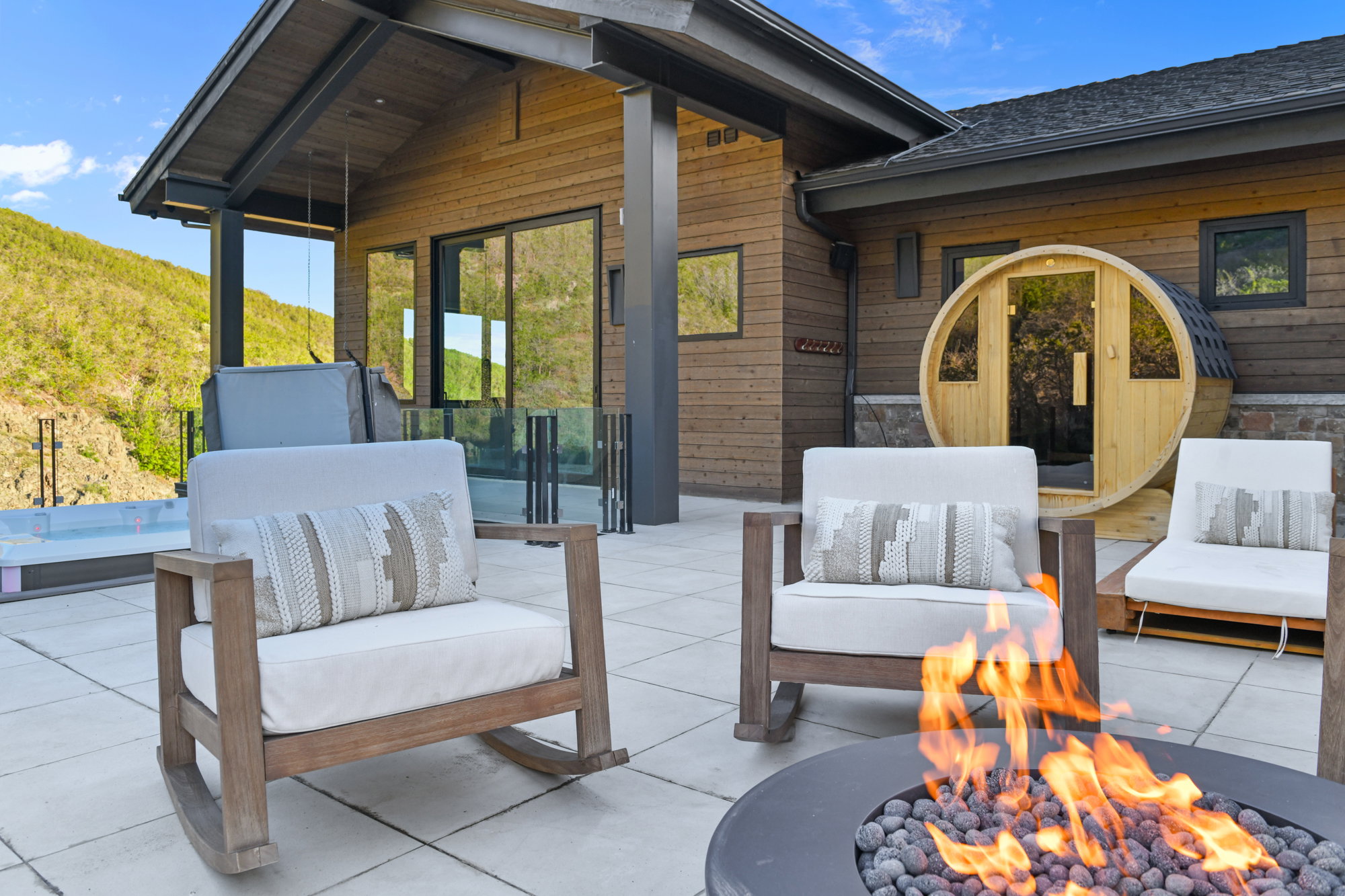 Expansive outdoor patio featuring a fire pit with stone fill, a wood-sided barrel sauna, and a glass-paneled hot tub