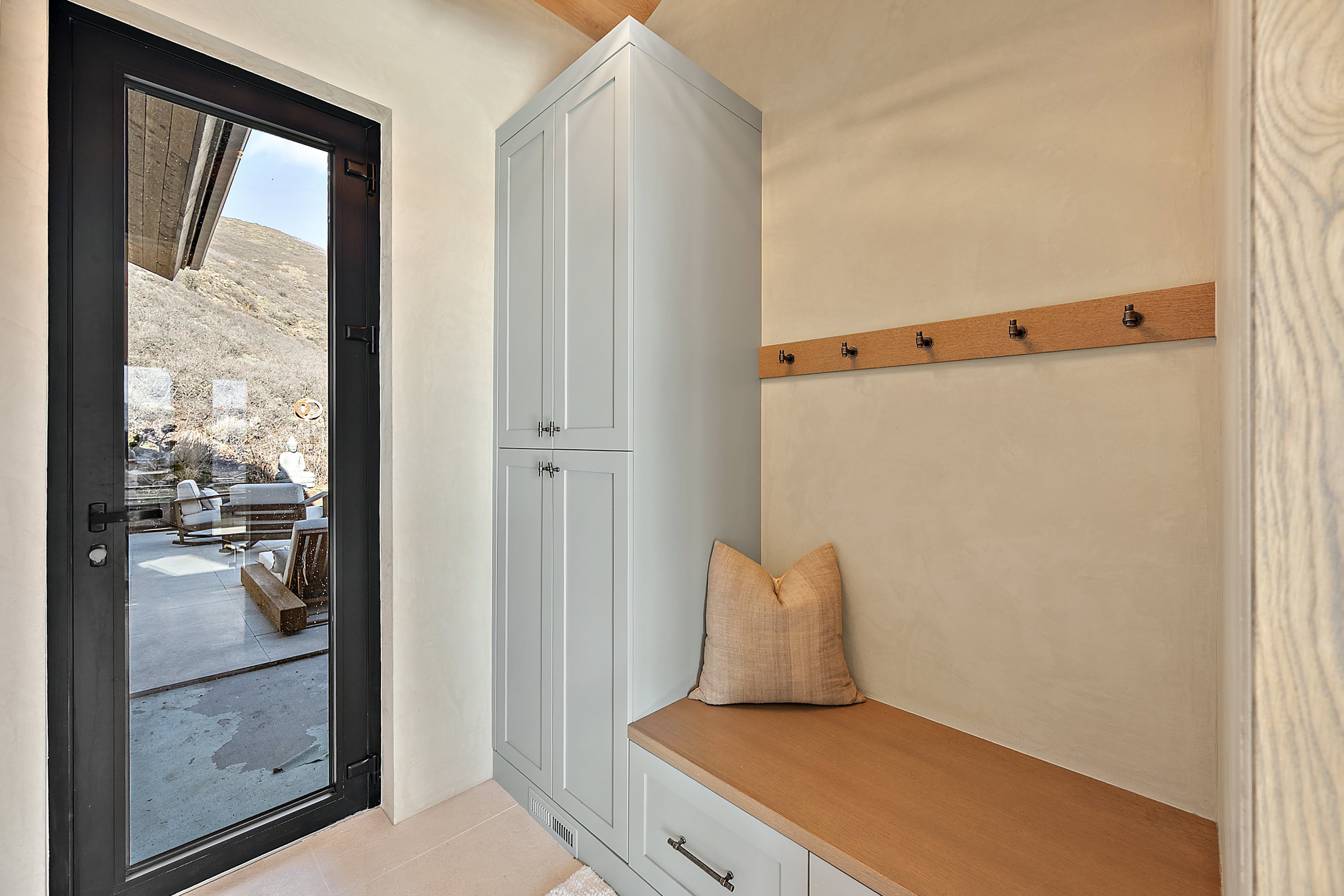 Full-light exterior door with black framing, built-in storage bench with wood-finish top, integrated cabinetry with shaker-style doors, and a wall-mounted coat rack