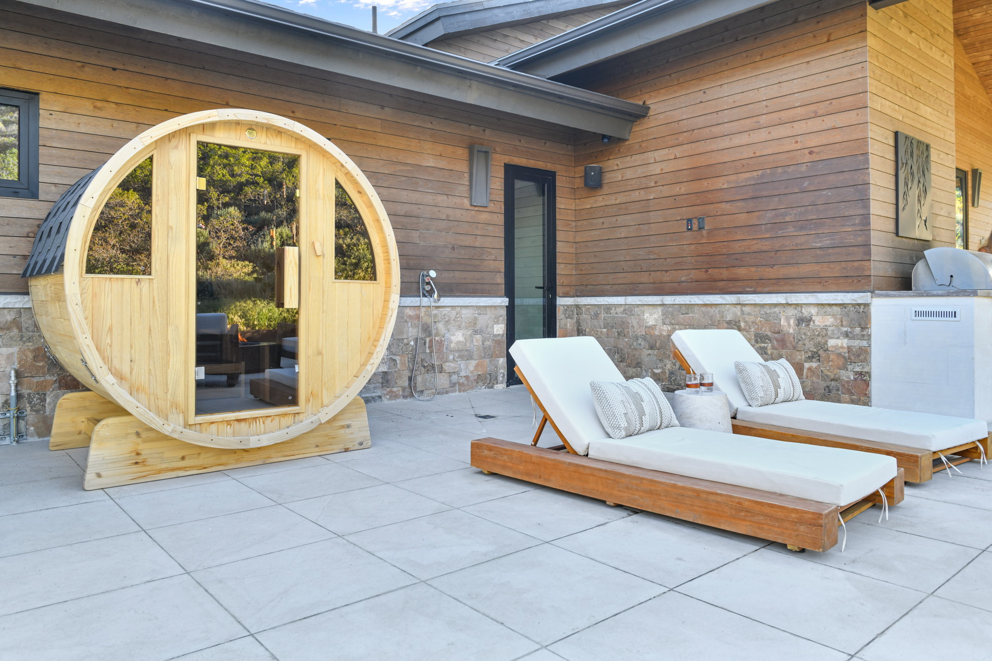 Outdoor barrel sauna with a shingled roof and glass door access, set on a tiled patio