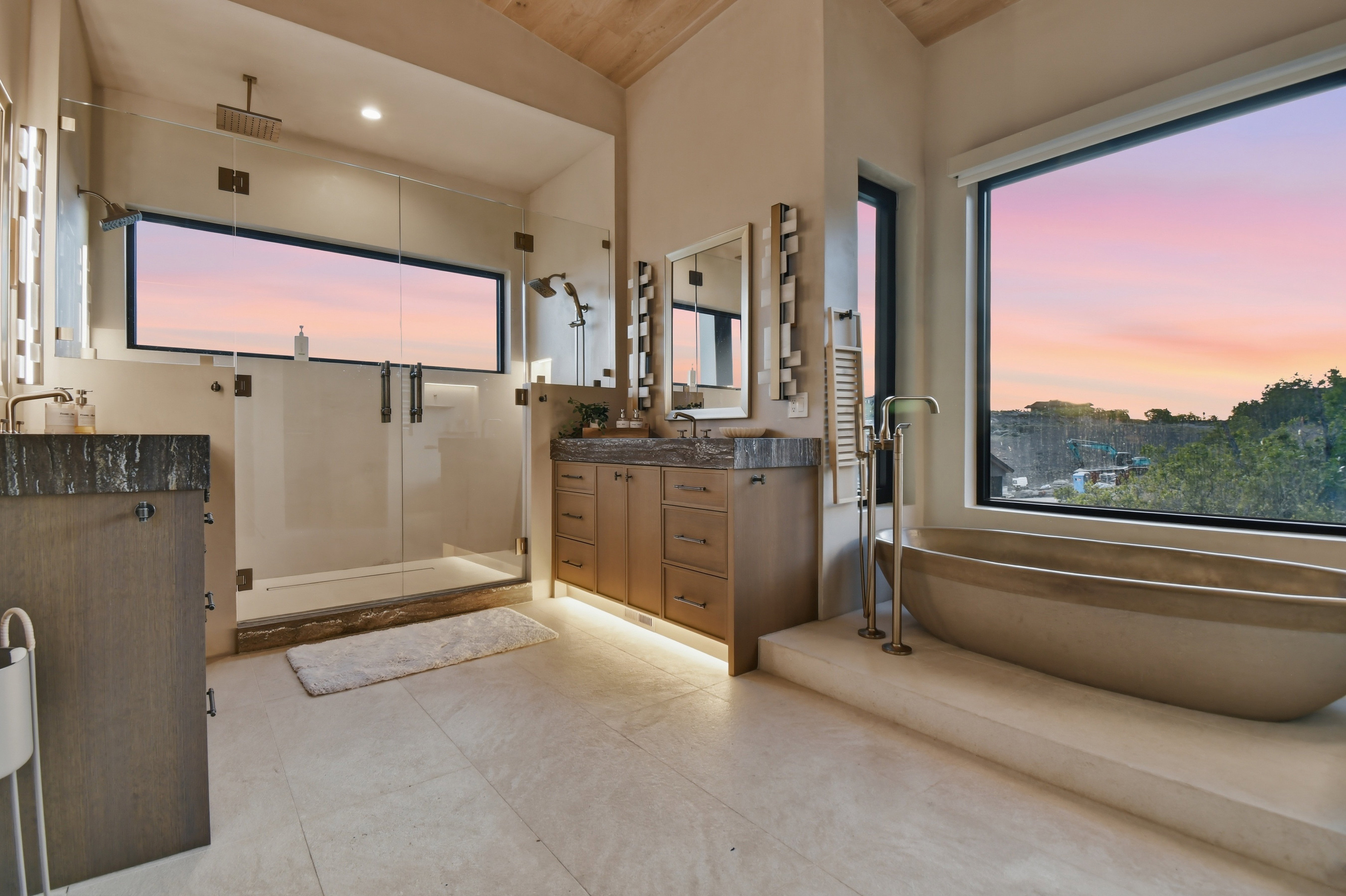 Spacious bathroom featuring a glass-enclosed shower with multiple showerheads, a freestanding soaking tub, twin wood vanities with stone countertops, and large windows