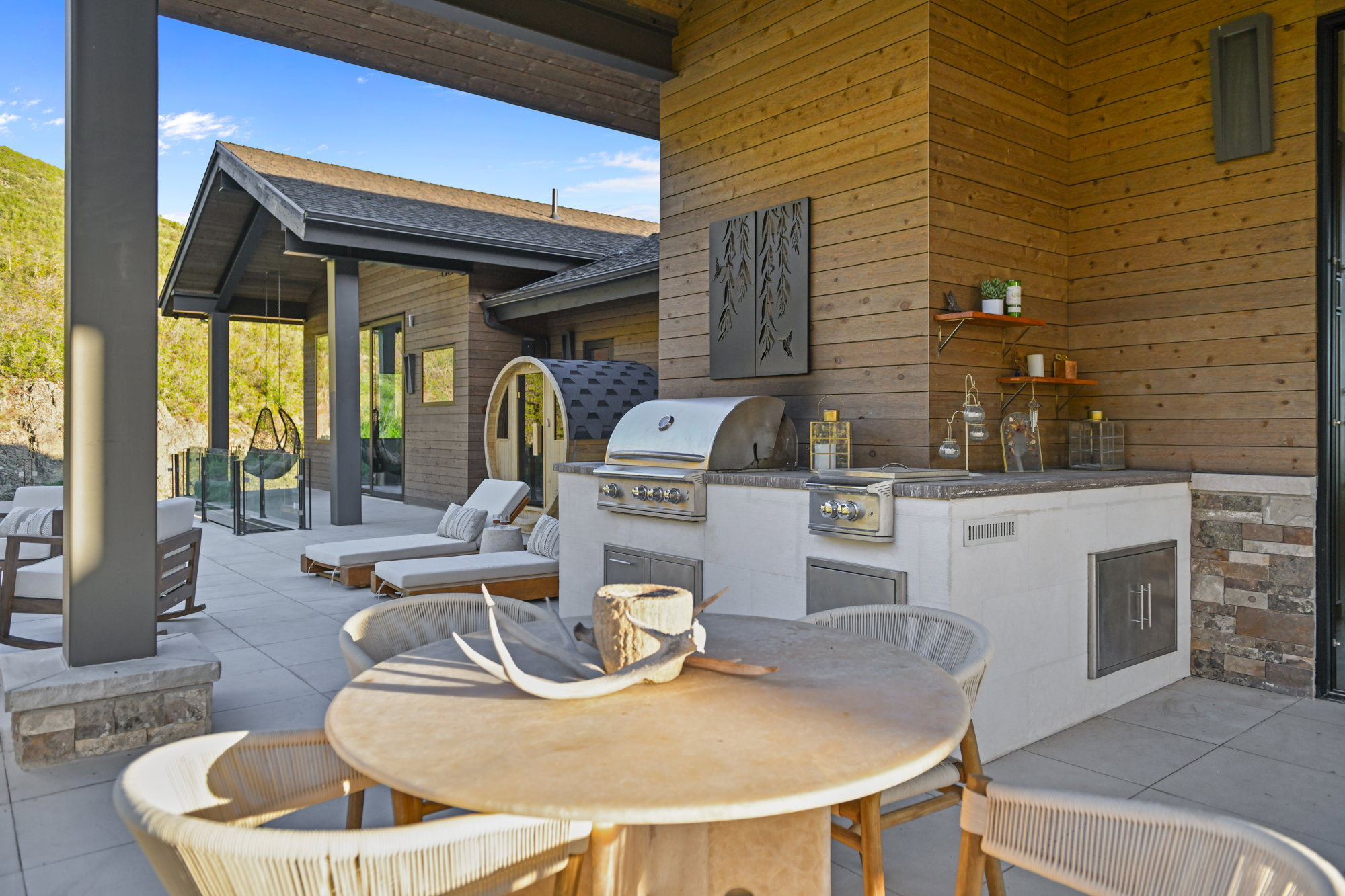 Expansive outdoor living area featuring a built-in outdoor kitchen with grill, wood-finish siding, a covered patio with recessed lighting, a stone base for structural columns, and a glass privacy barrier
