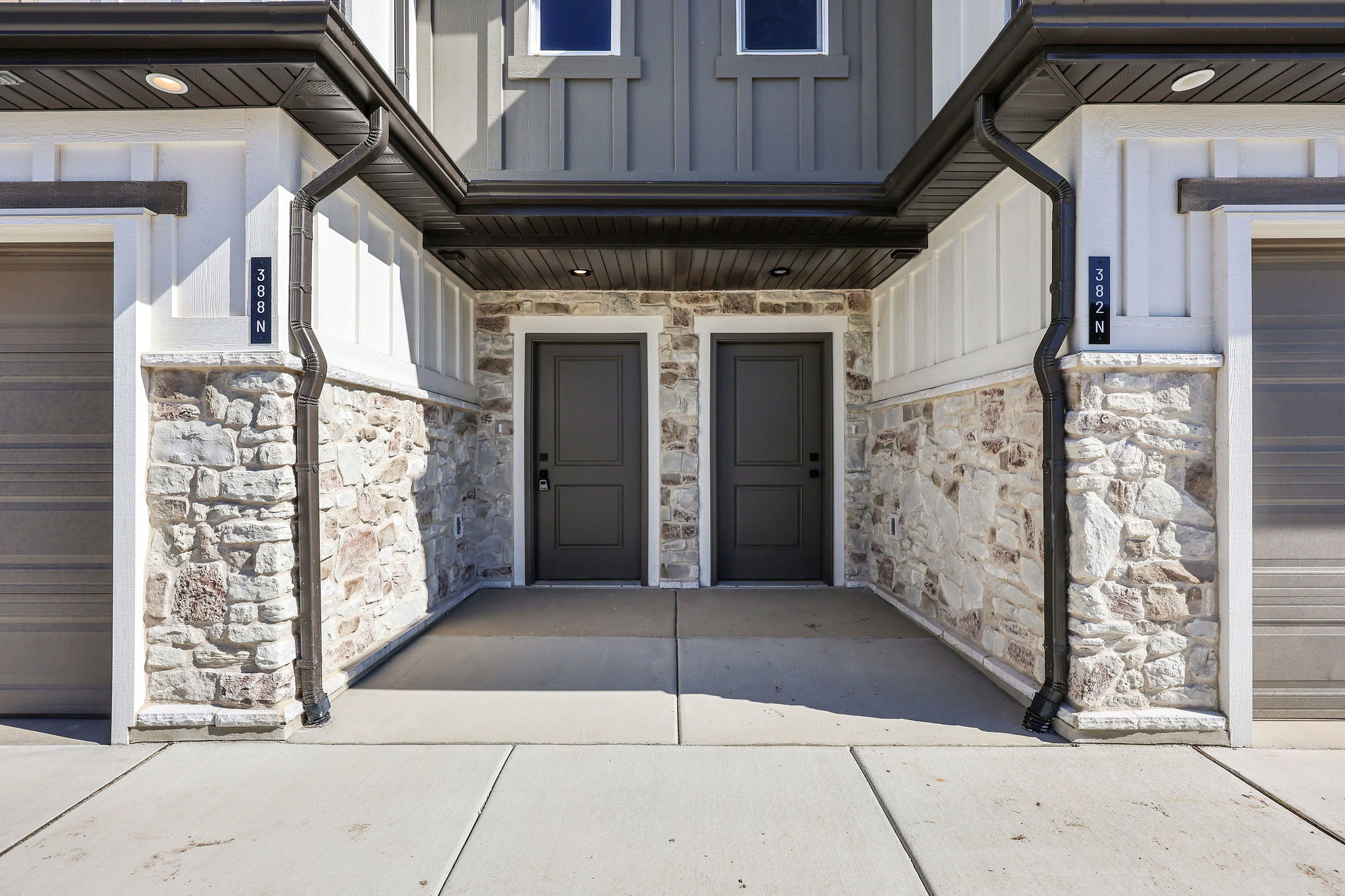 Modern townhome exterior featuring a stone facade, dark wood-finish garage doors, dark entry doors, white board and batten siding, and dark gutters
