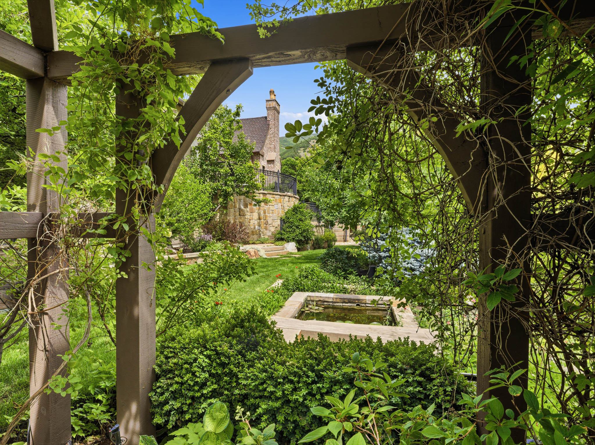 Lush backyard landscaping featuring a wooden pergola with climbing foliage, a central water feature, manicured lawns, mature trees, and a stone retaining wall