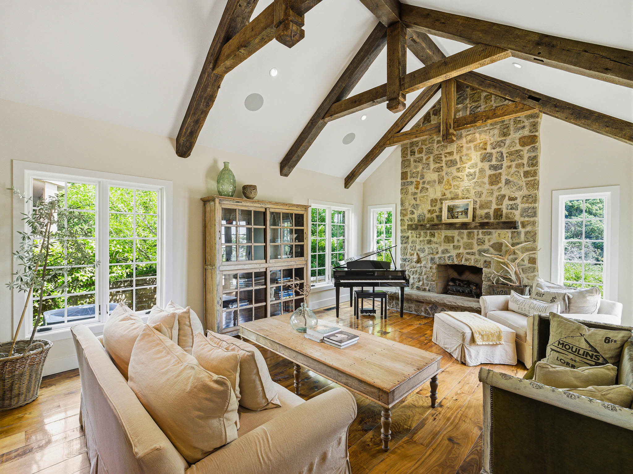 Spacious living area featuring a vaulted ceiling with exposed wood beams, a stone-clad fireplace, and multiple grid-pattern windows