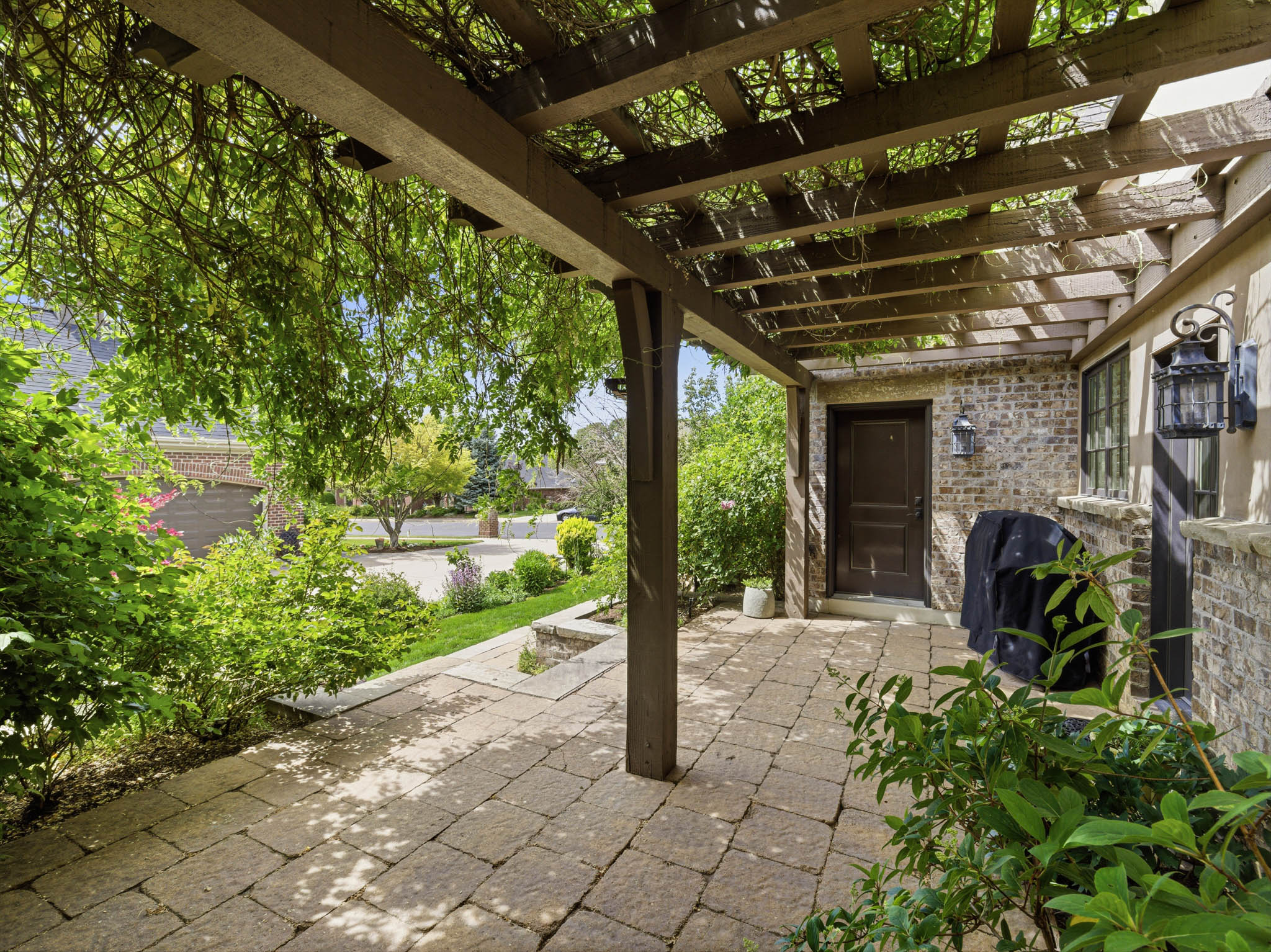 Covered patio featuring an open-beam pergola with leafy vine coverage, brick exterior accents, and paver stone flooring