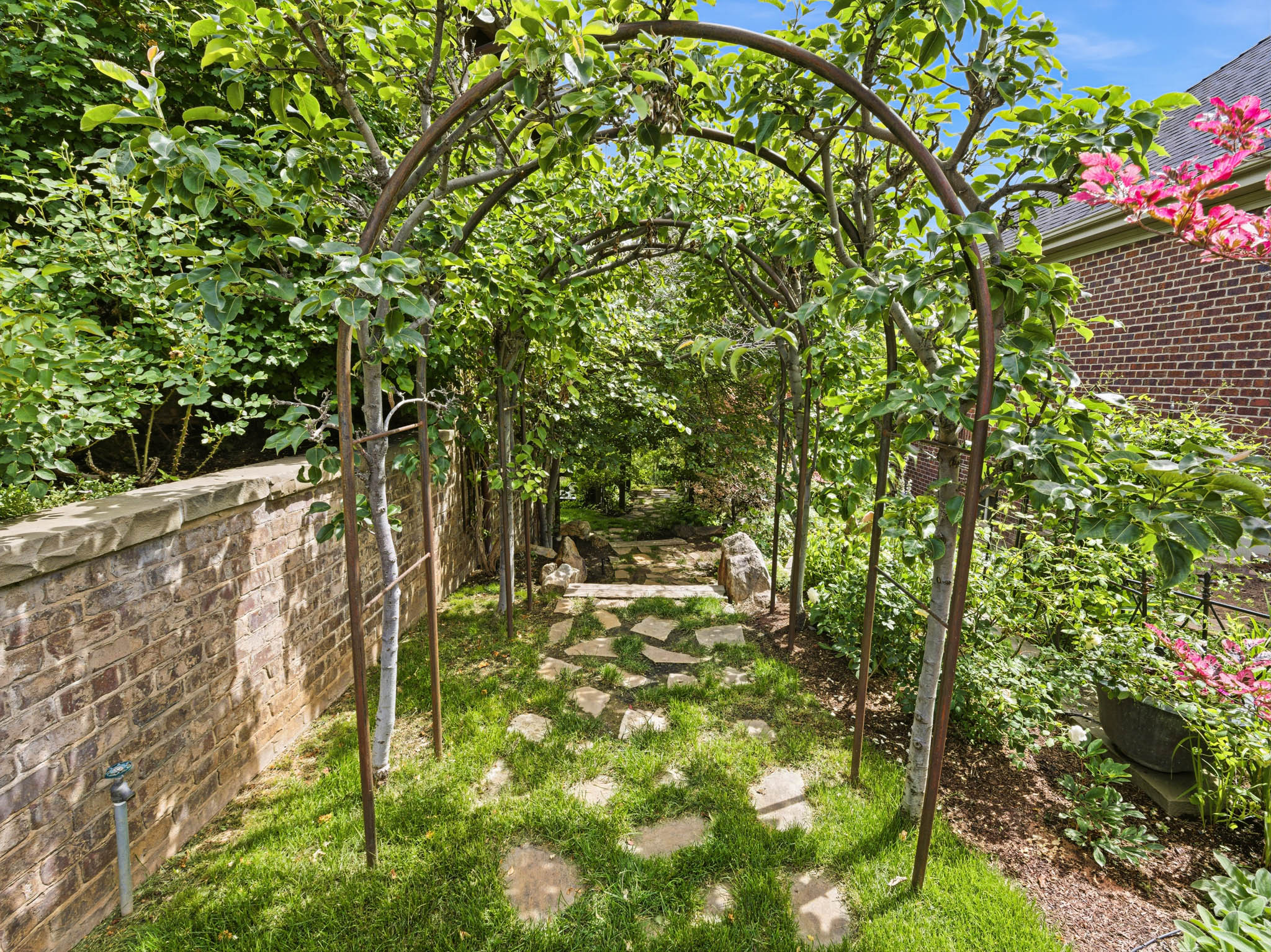 Landscaped garden pathway featuring a mature arbor with lush greenery, flagstone steps, manicured grass, a brick retaining wall, and verdant plantings