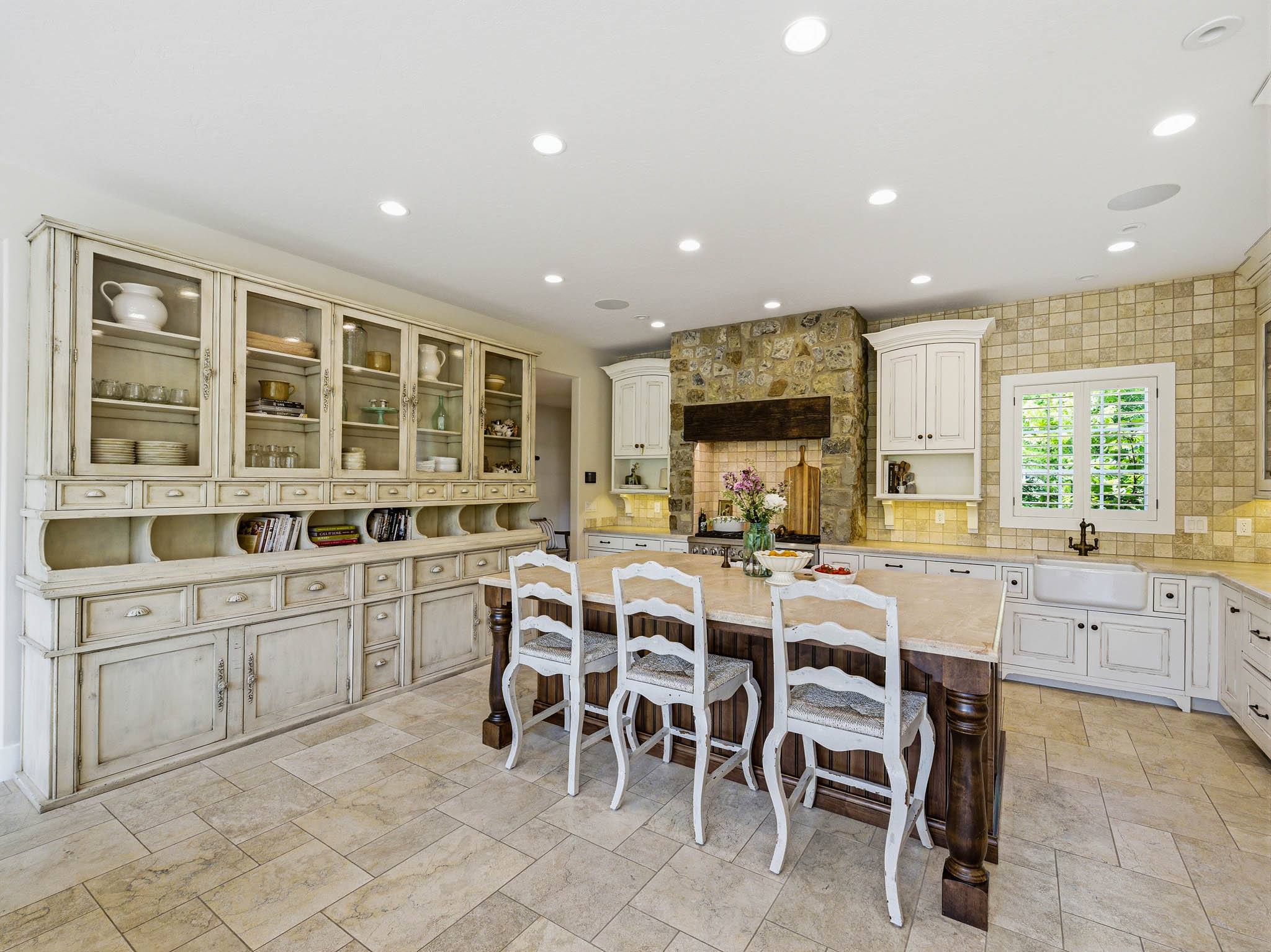 Spacious kitchen featuring a large central island with a wood base and light-toned countertop, extensive built-in cabinetry with glass display doors and drawers, stone tile flooring, a stone-clad fireplace with a wood mantel, and a farmhouse sink with a d