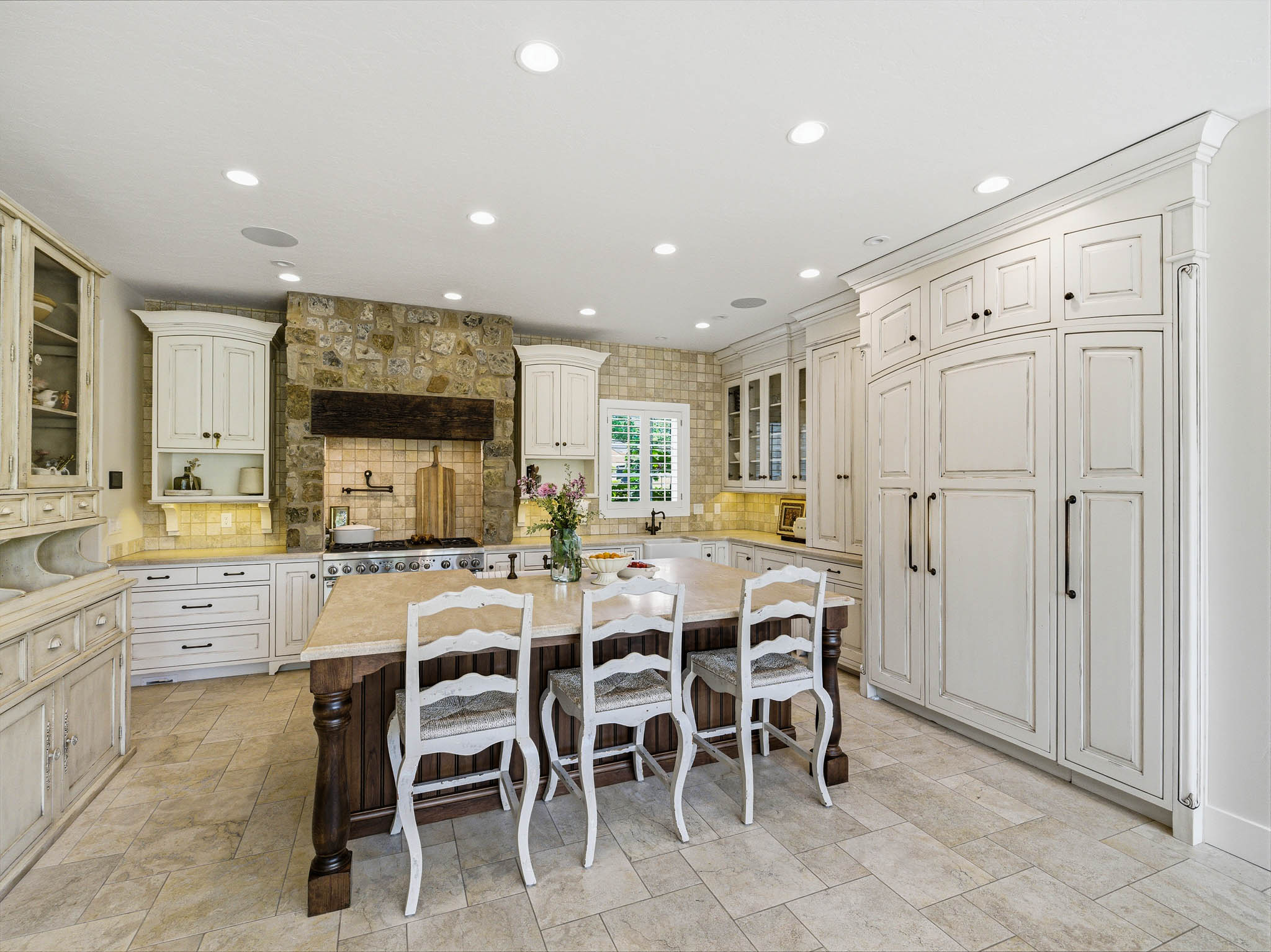 Spacious kitchen featuring a large central island with a light-toned countertop, extensive white cabinetry with dark hardware, a stone-clad range hood and backsplash, professional-grade range, and recessed lighting
