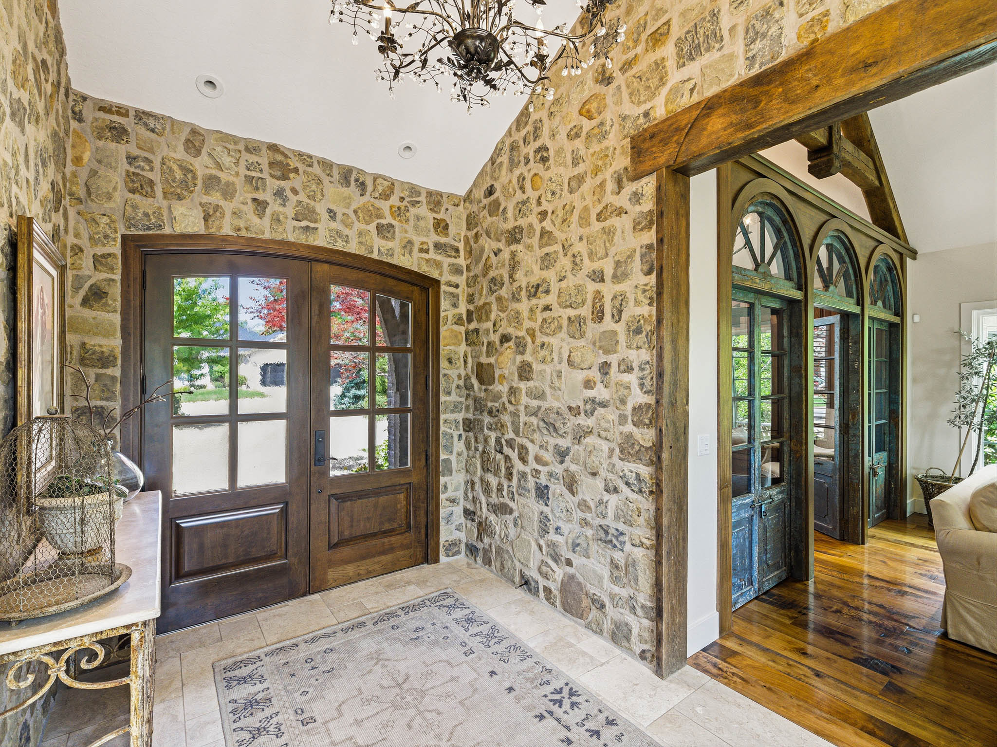Foyer featuring stone walls, arched wood-frame glass panel double doors, a decorative chandelier, tile flooring, and a wood beam archway