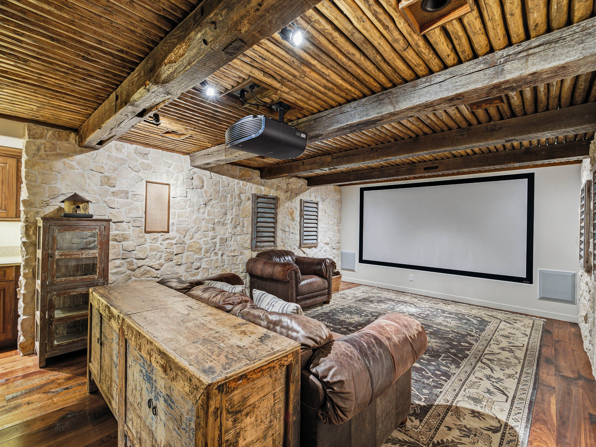 Media room featuring a large projection screen, stone accent walls, exposed wood beam ceiling, wood-finish flooring, and integrated audio speakers