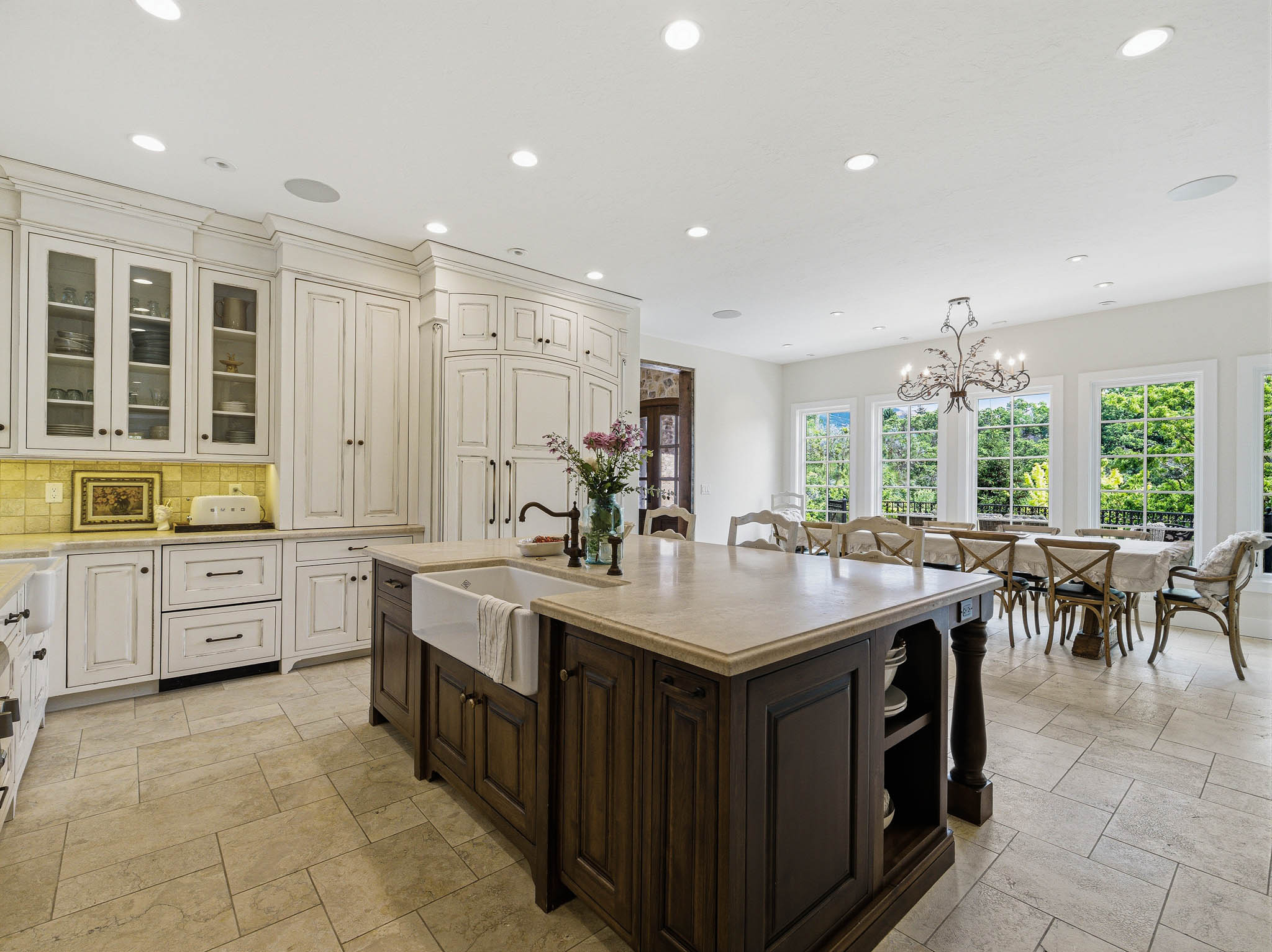 Spacious kitchen featuring a central island with an integrated farmhouse sink, extensive cabinetry with glass-front uppers, recessed ceiling lighting, and tile flooring, opening to a dining area with multiple picture windows