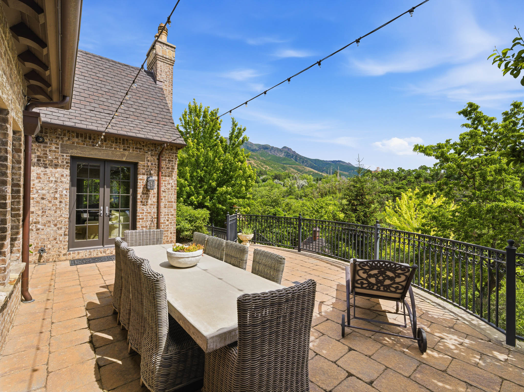 Expansive outdoor patio featuring paver stone flooring, a decorative wrought iron railing, a brick exterior facade, and a prominent chimney structure