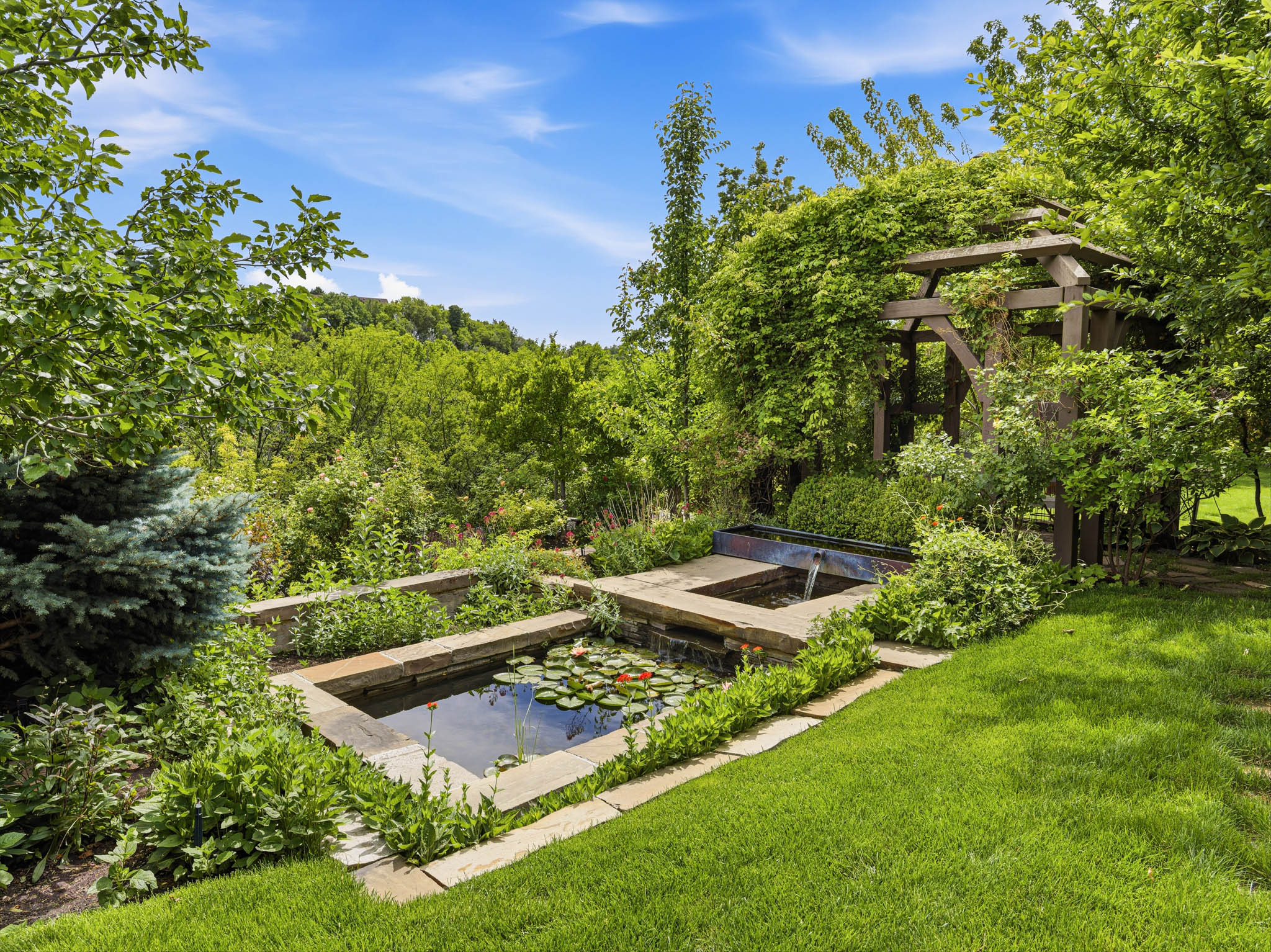Expansive backyard featuring a multi-tiered water feature with a koi pond, surrounding stone coping, a cascading water element, a wooden pergola covered in climbing foliage, and a lush green lawn
