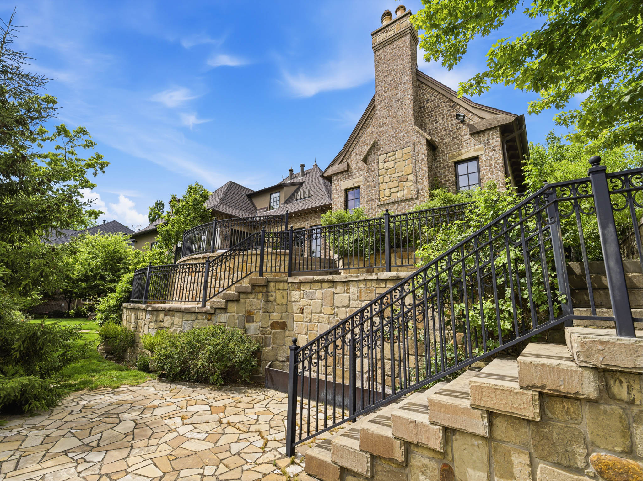 Stone and brick exterior with a prominent brick chimney, multi-level stone hardscaping, wrought iron railings, a stone patio, and dormer windows