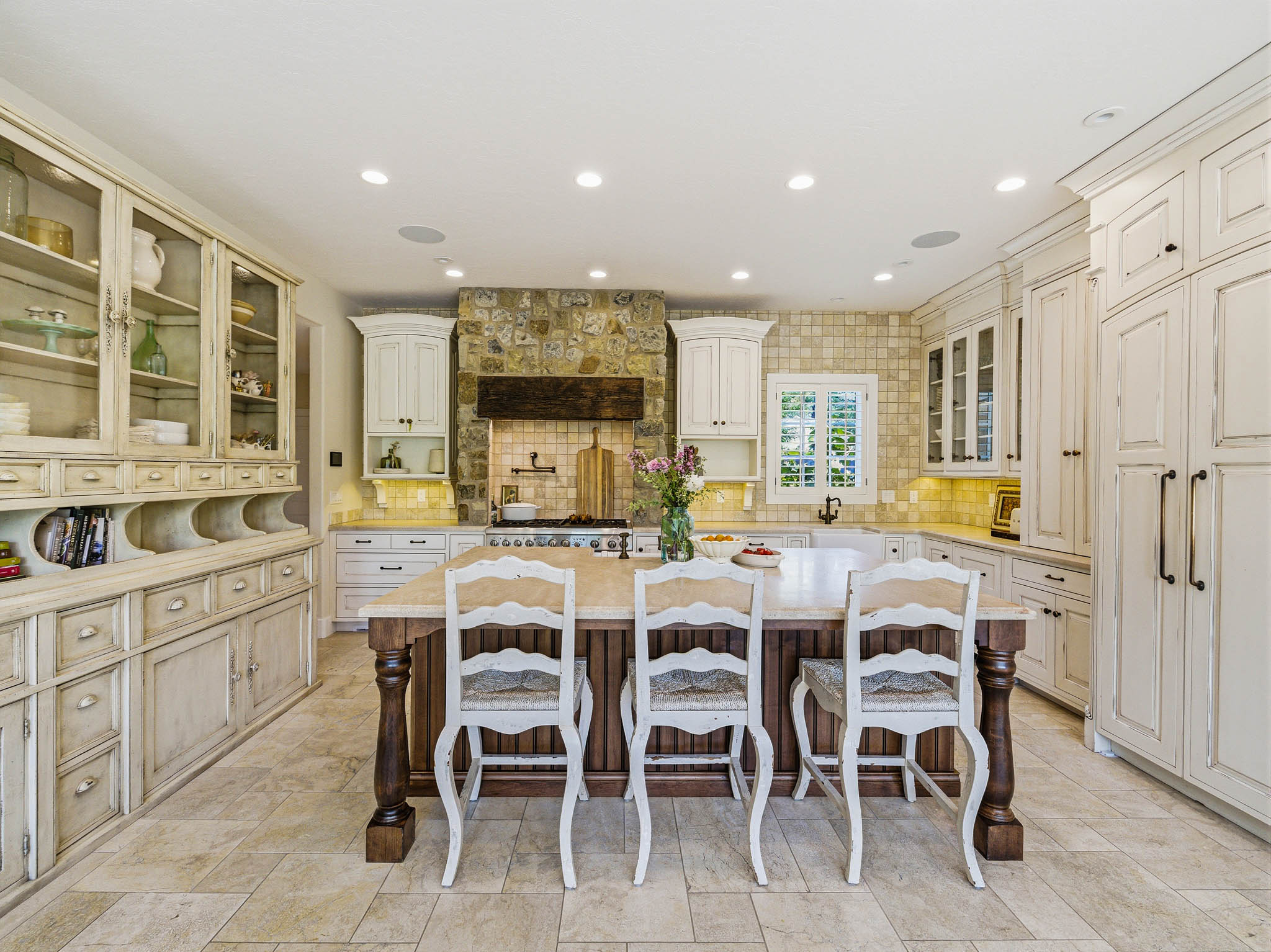 Kitchen featuring a central island with a light-toned countertop, extensive built-in cabinetry with glass-front uppers, a stone-clad range hood, tile flooring, and recessed lighting
