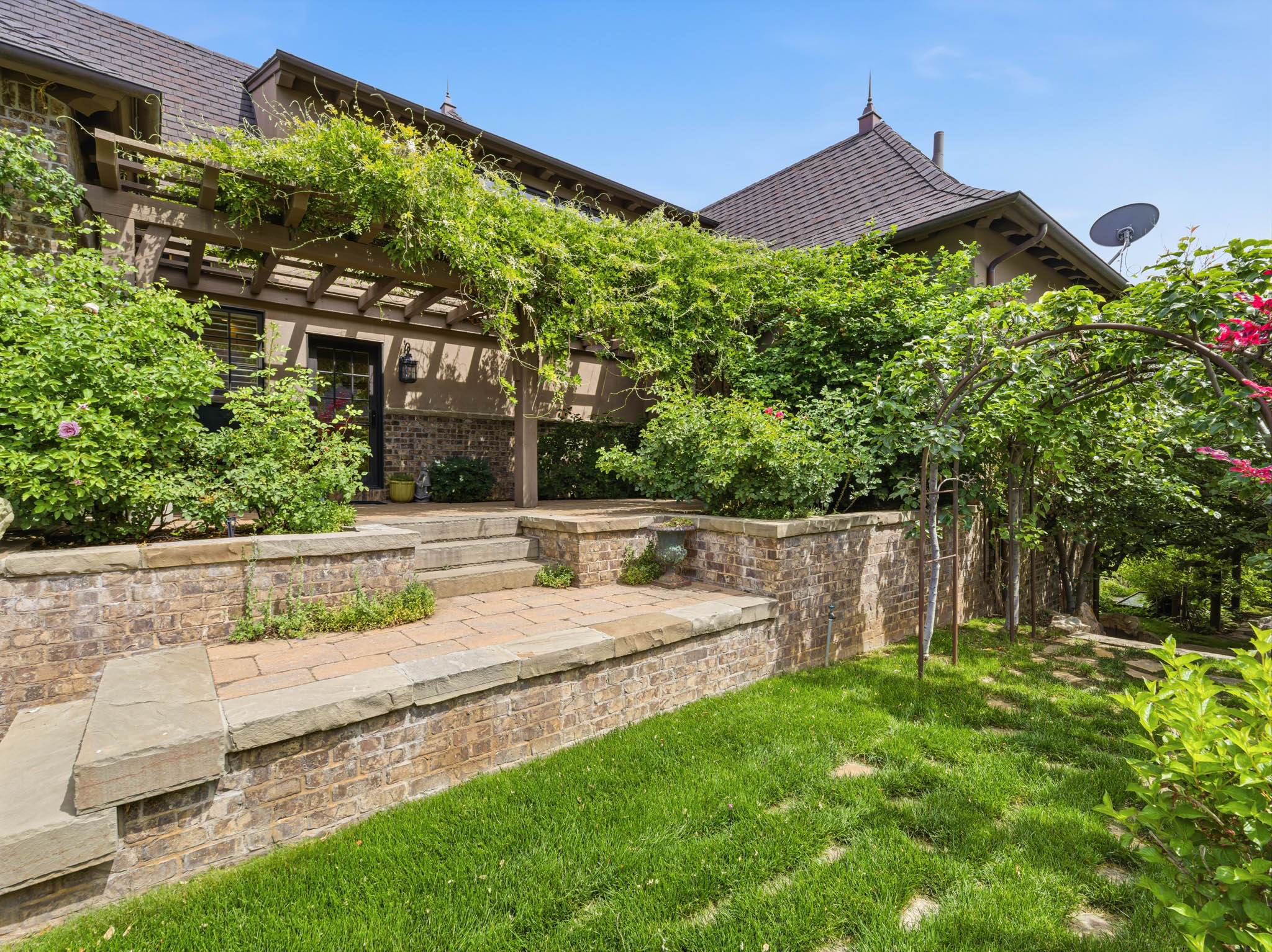 Terraced exterior landscaping featuring brick retaining walls with stone caps, stone paver patio, wood pergola with abundant vines, and a dark tile roof