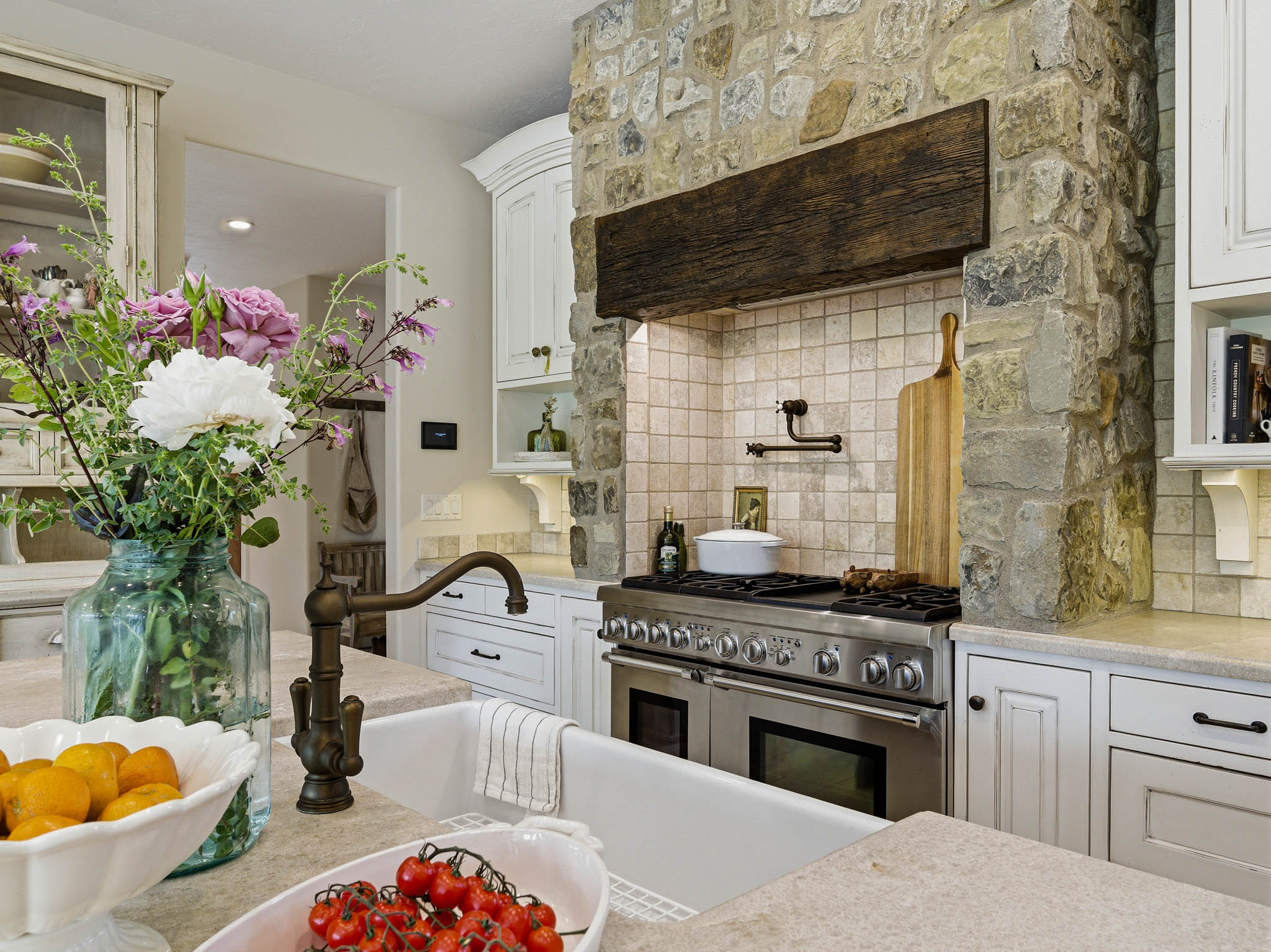 Gourmet kitchen featuring a professional-grade stainless steel range, built-in stone hood with wood-finish beam, tile backsplash, and a farmhouse sink with a bronze-finish faucet