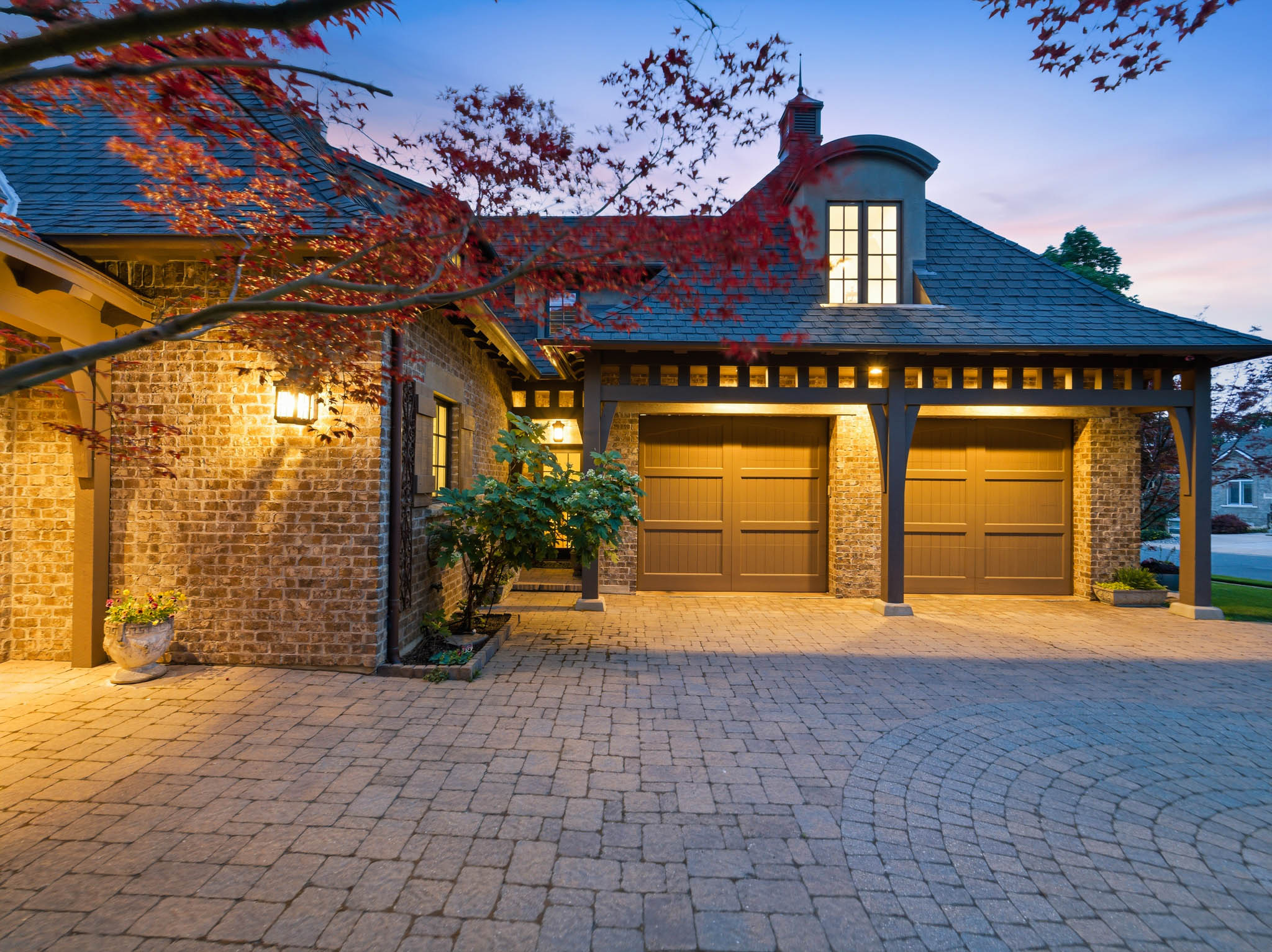 Brick exterior featuring a multi-car garage with wood-paneled doors, a curved dormer, a slate-style roof, and a paver driveway with a circular inlay