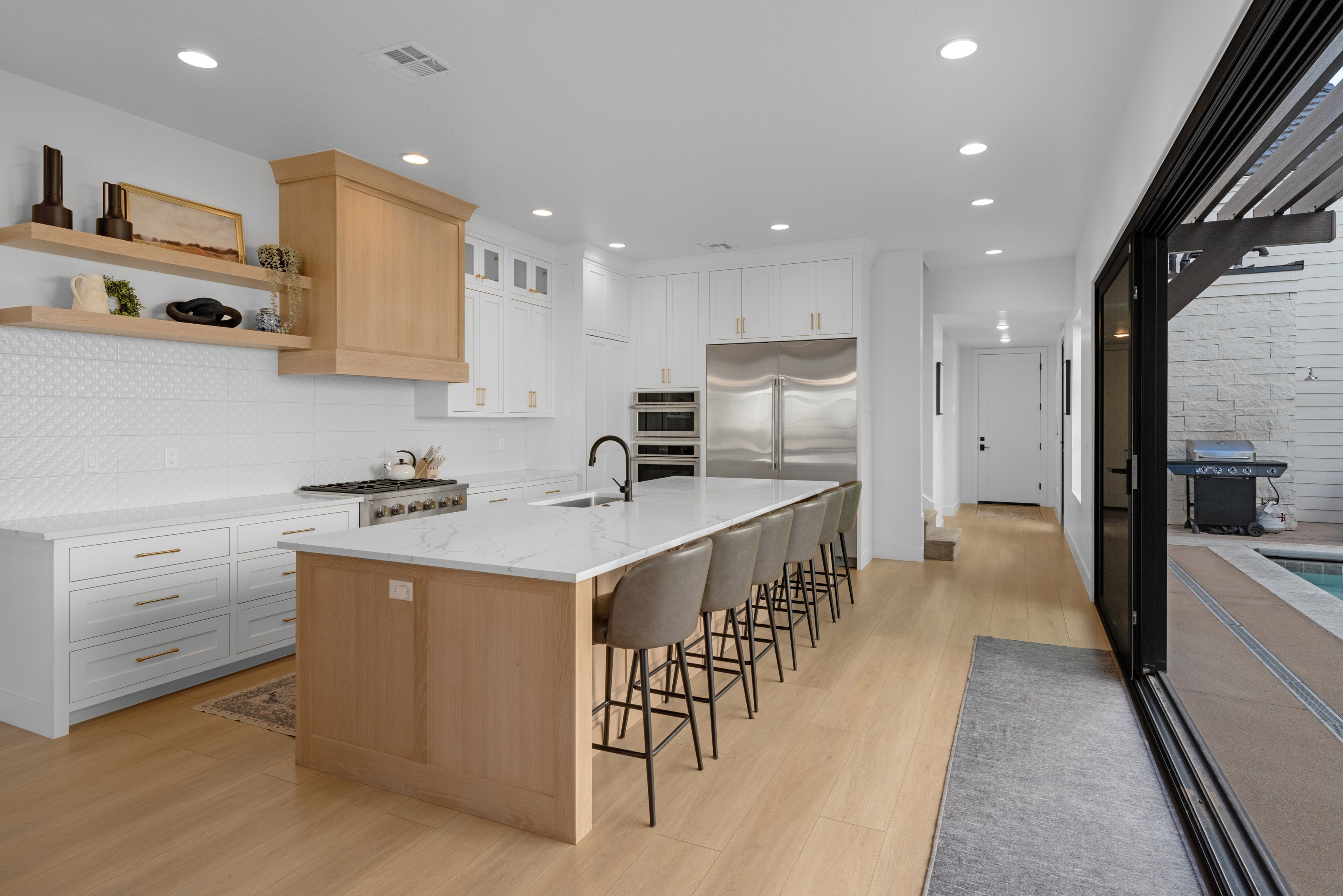 Gourmet kitchen featuring a large central island with seating, stainless steel appliances, white cabinetry with gold-tone hardware, a wood-finish range hood, and light wood-finish flooring