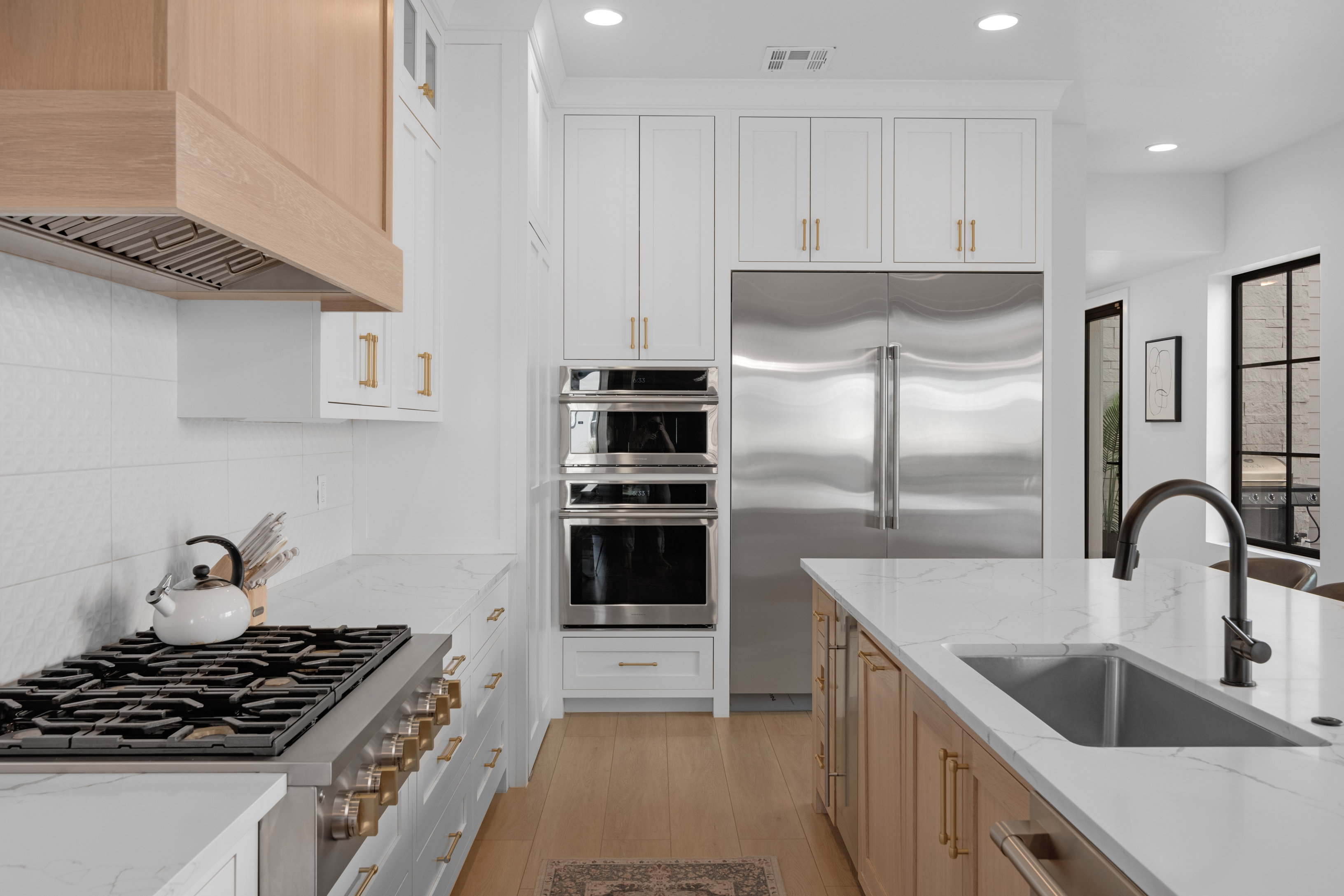 Gourmet kitchen featuring a built-in stainless steel refrigerator, double wall ovens, a large gas range with a wood-finish vent hood, a spacious island with an undermount sink and matte black faucet, and light wood-finish flooring