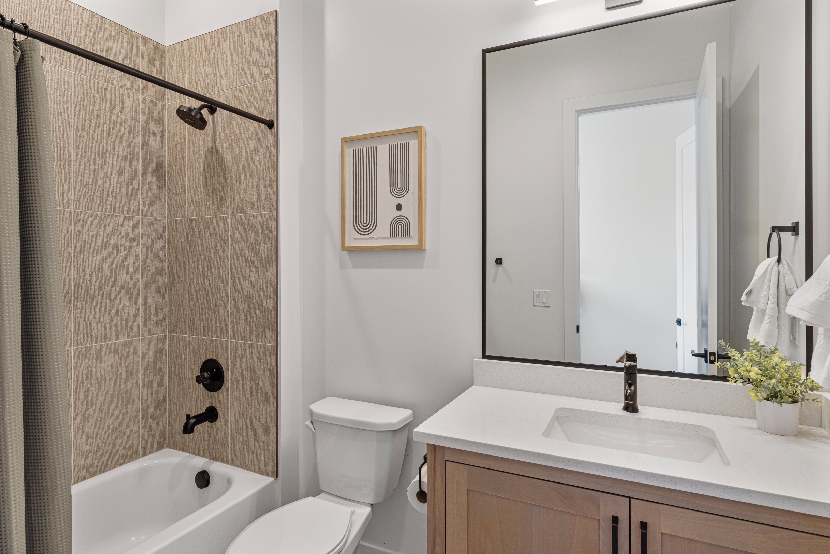 Bathroom featuring a light wood-finish vanity with a white countertop and integrated sink, a rectangular framed mirror, and a shower-over-tub combination with large format tiled walls and matte black fixtures