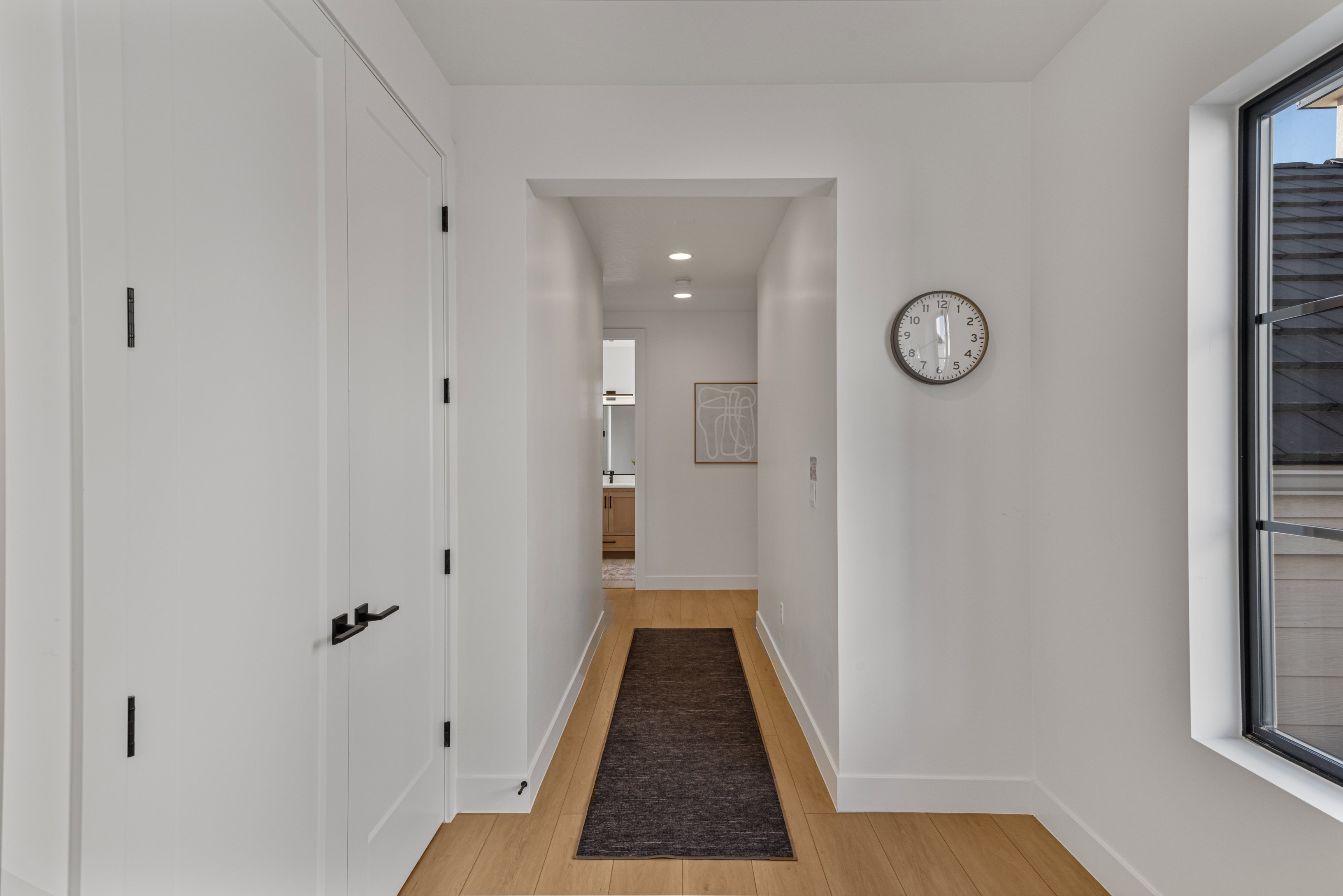 Bright hallway featuring light wood-finish flooring, white walls, recessed lighting, and a window with black trim