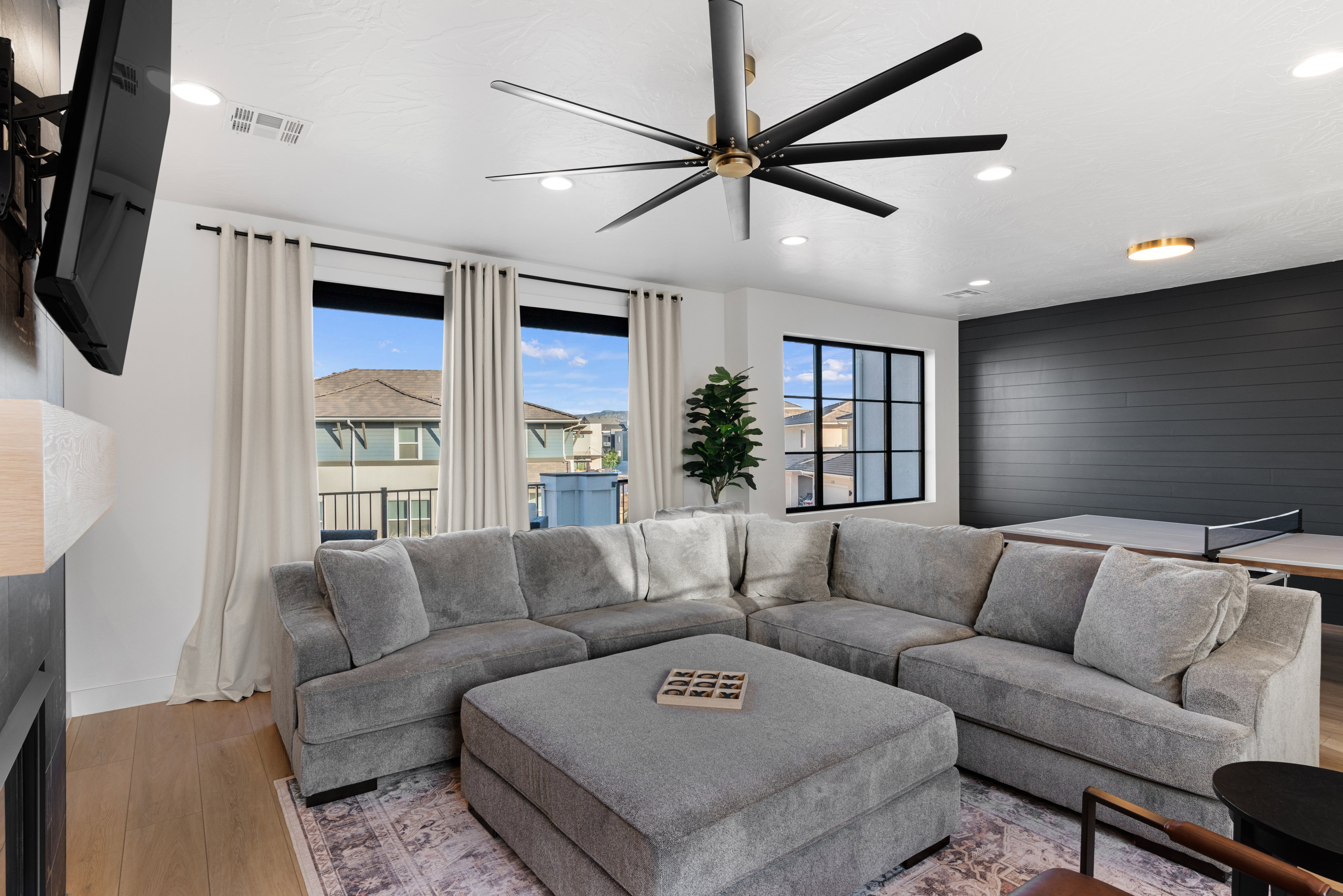 Spacious living area featuring light wood-finish flooring, a contemporary ceiling fan, recessed lighting, and large windows with privacy curtains