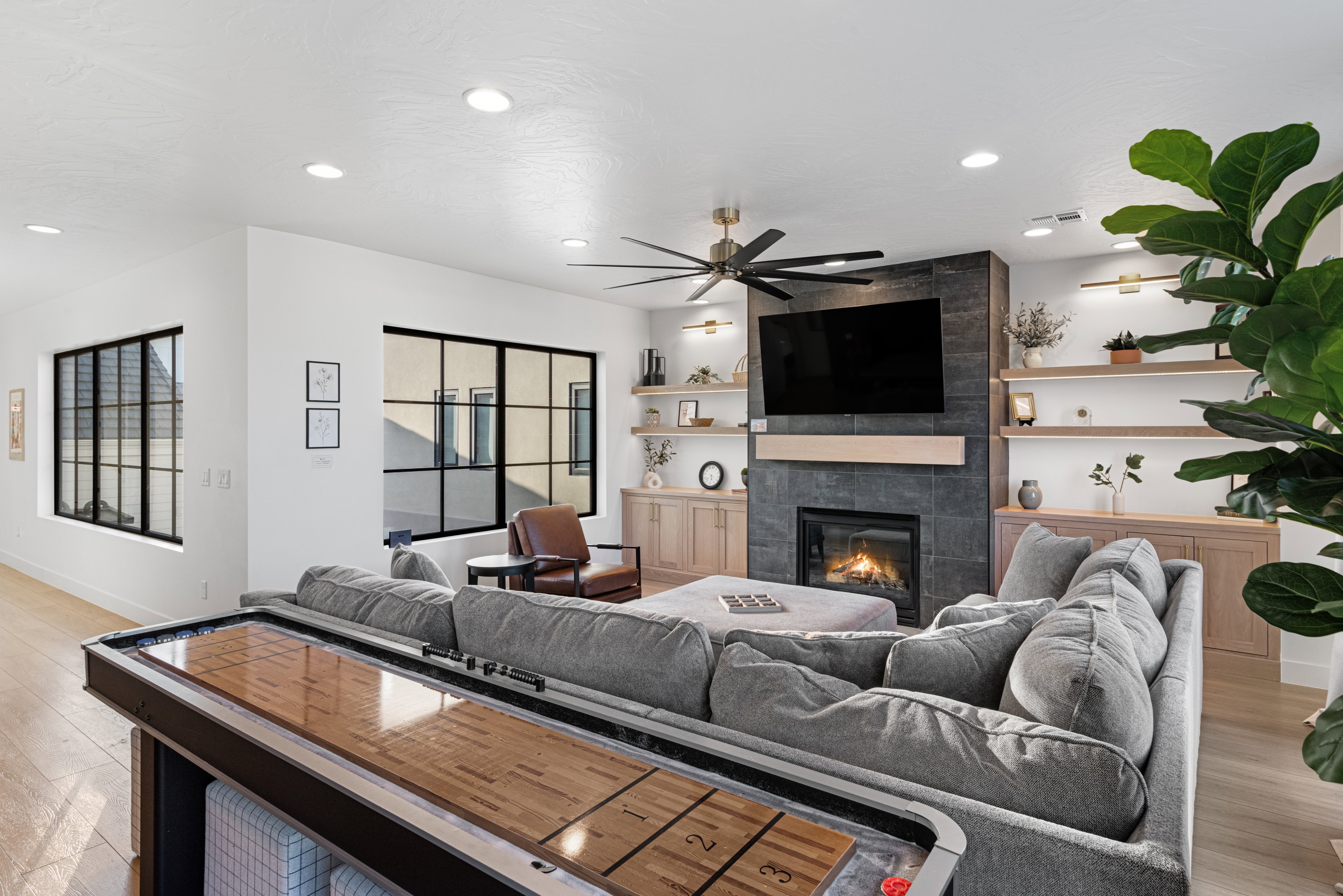 Spacious living area featuring a stone-clad fireplace, integrated wood-finish shelving and cabinetry, recessed ceiling lighting, a contemporary ceiling fan, and black-framed grid windows