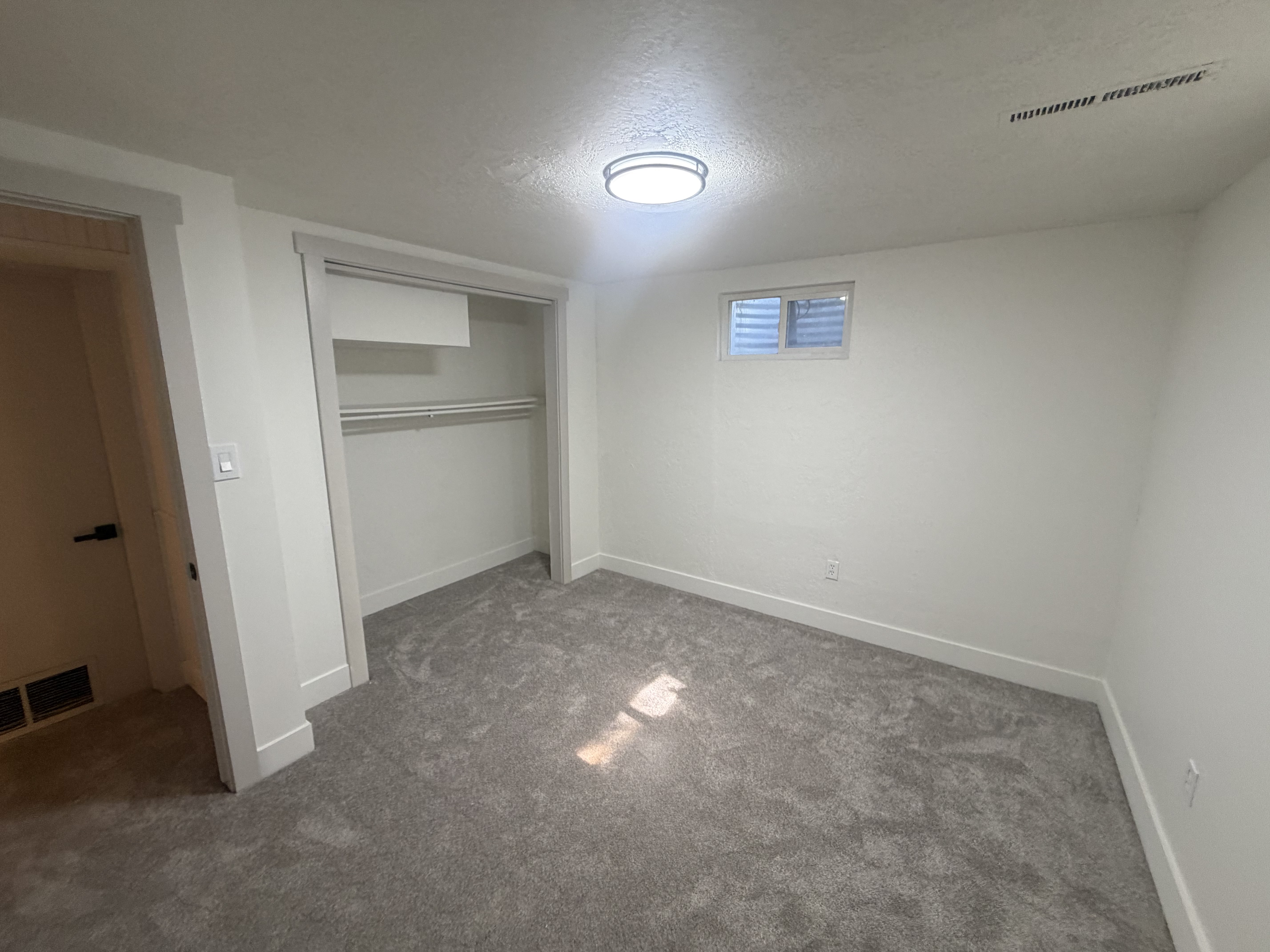 Bedroom 5. Basement. Carpeted room featuring a ceiling-mounted light fixture, a built-in closet with shelving and hanging rods, a single window, and a white door with a black handle