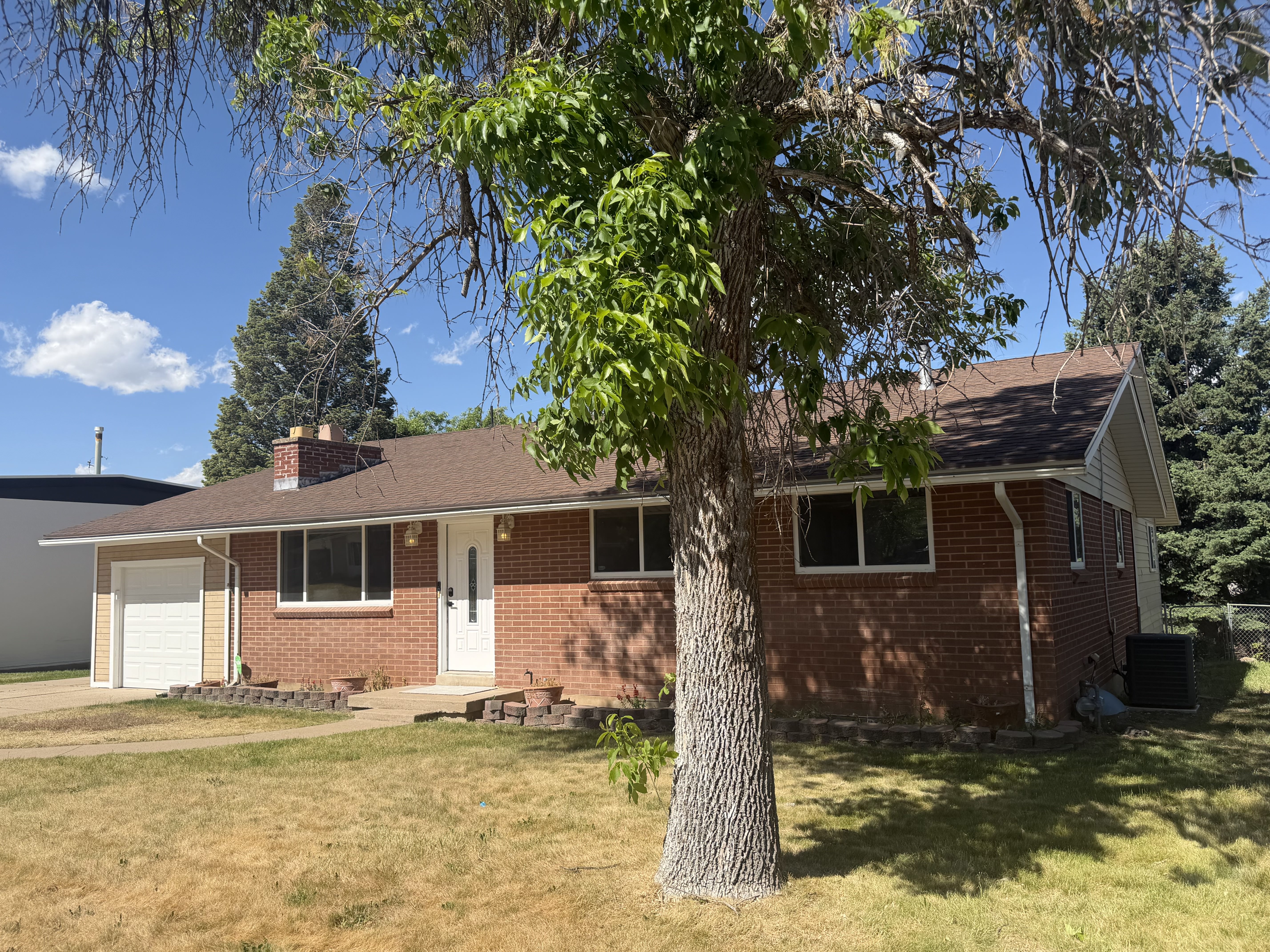 Brick ranch-style residence featuring a single-car garage with a white door, a white front entry door, a brick chimney, and a brown shingle roof