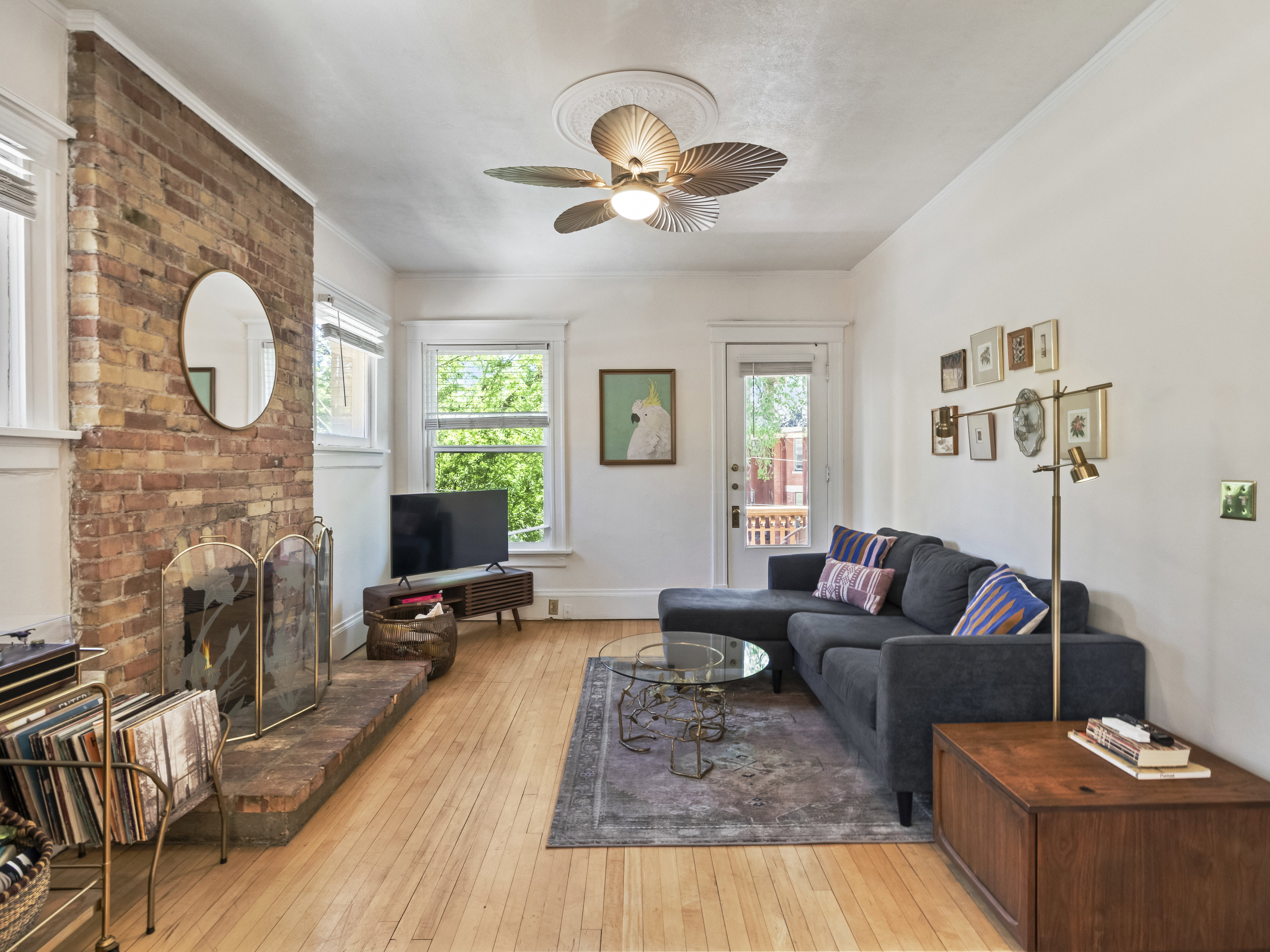 Exposed brick fireplace with hearth, wood-finish flooring, white trim windows, ceiling fan with light fixture, and a glass-paneled door