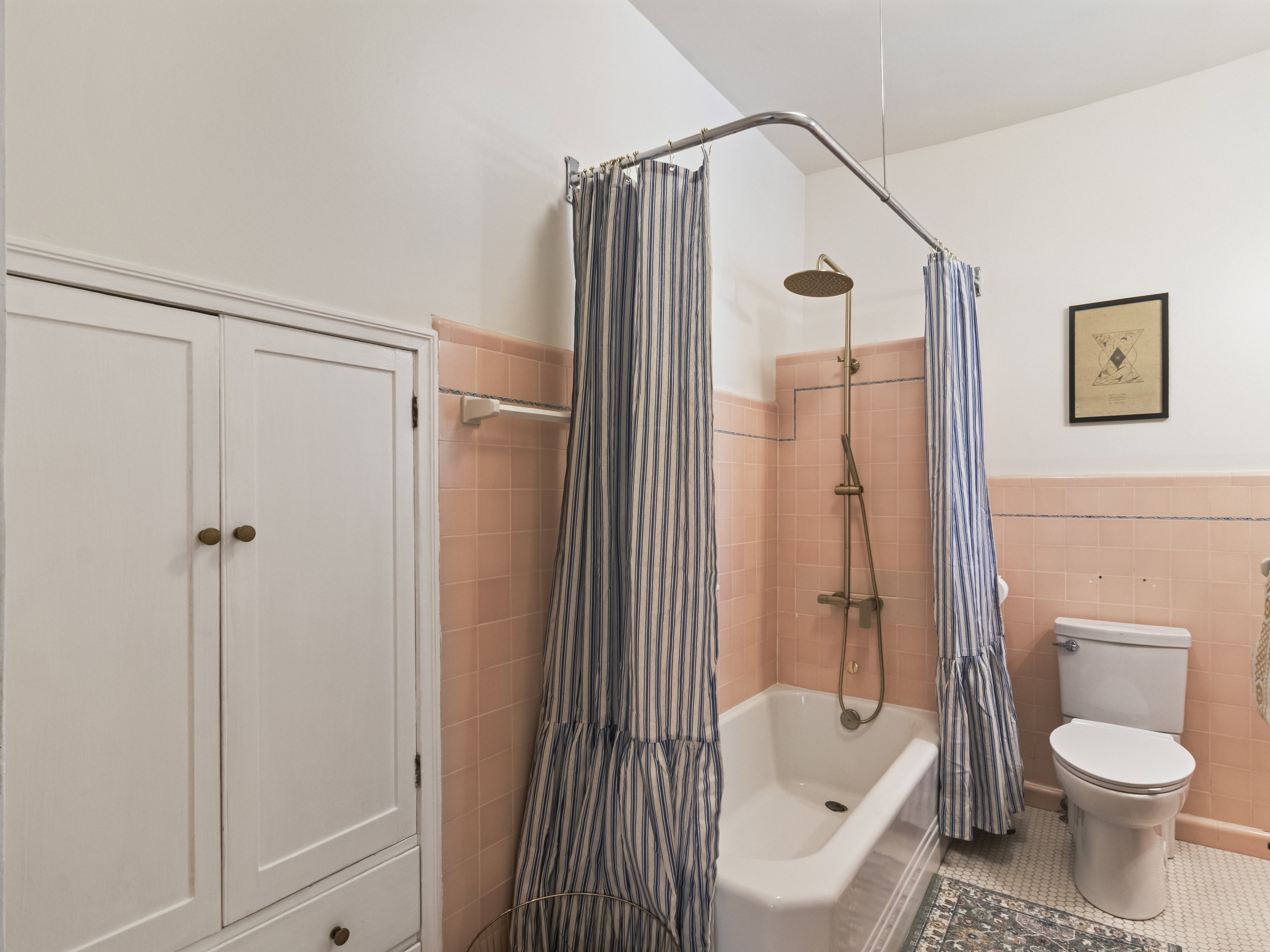 Bathroom featuring a clawfoot tub with an overhead shower fixture, pink tiled walls, a white toilet, and white built-in cabinetry with wood knobs