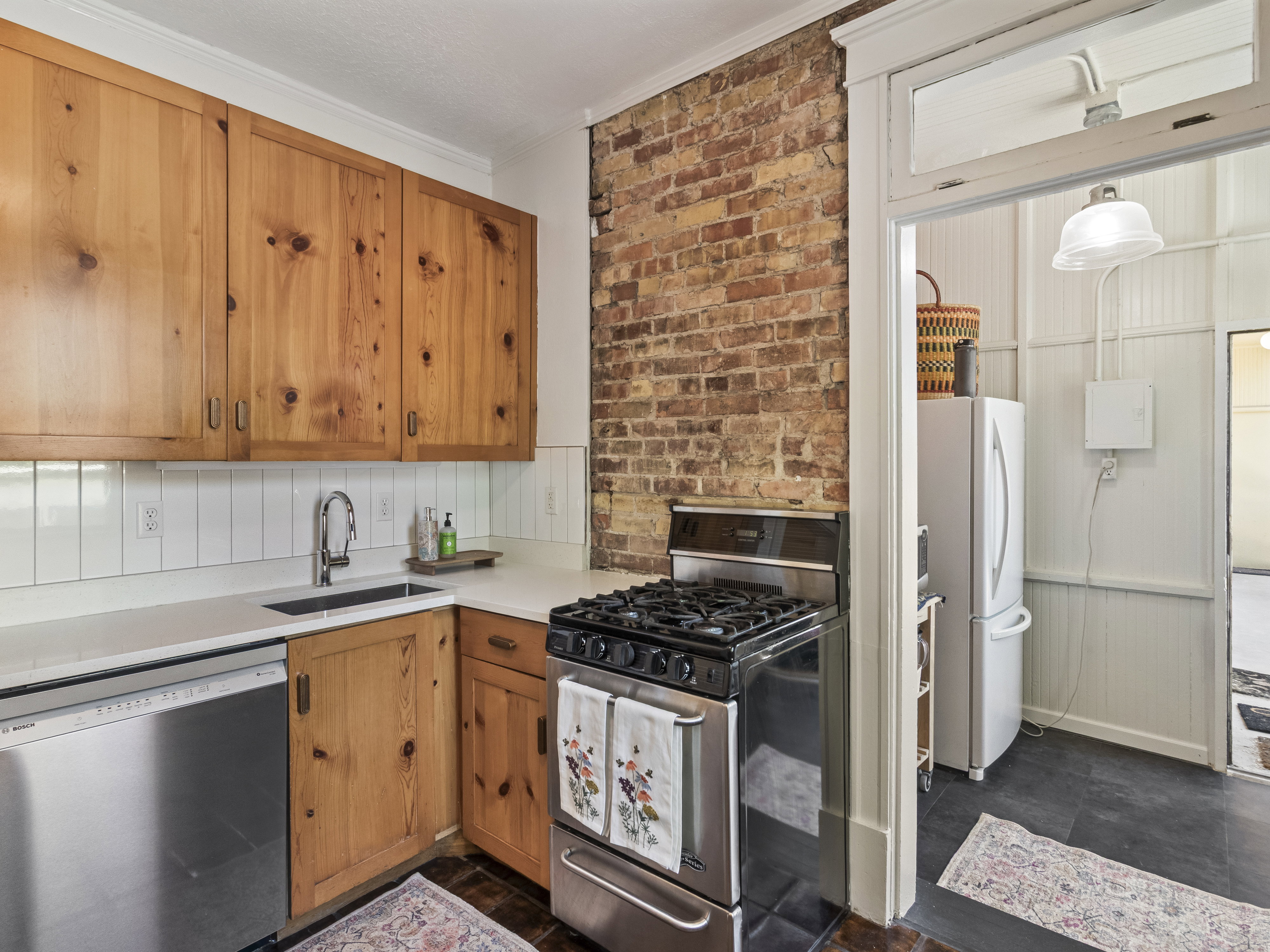 Kitchen featuring natural wood cabinetry, exposed brick accent wall, white subway tile backsplash, stainless steel gas range, and a built-in dishwasher