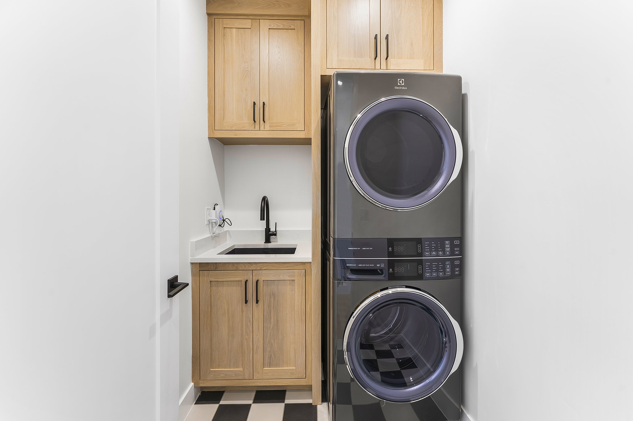 Dedicated laundry area featuring a stacked washer and dryer, light wood-finish cabinetry, a black sink with a matte black faucet, and a light-colored countertop