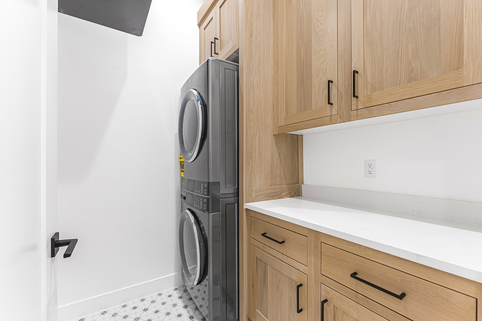 Laundry area featuring light wood cabinetry with matte black pulls, white countertop, white walls, and a patterned tile floor