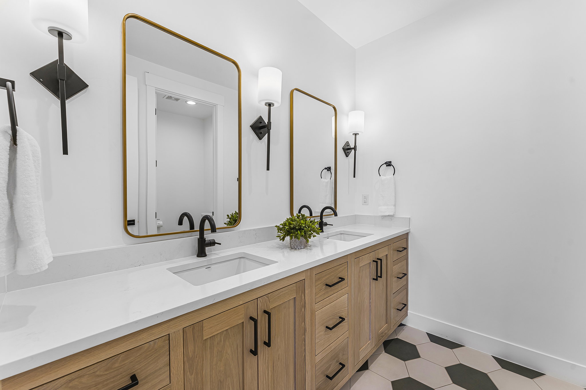 Dual vanity featuring light wood-finish cabinetry, white countertops, undermount sinks with matte black faucets, and hexagonal floor tile in contrasting tones