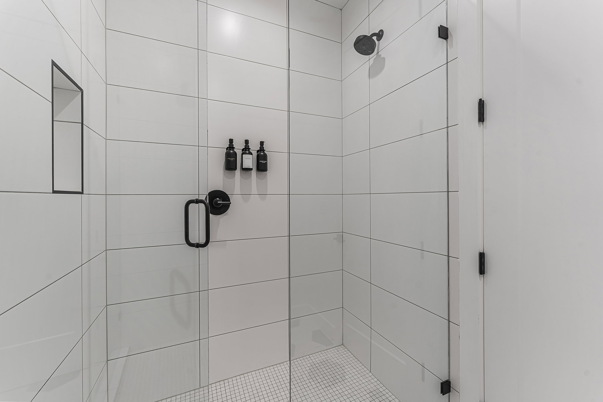 Shower enclosure featuring clear glass panels, matte black hardware, white rectangular wall tiling, a recessed niche, and a mosaic tile floor