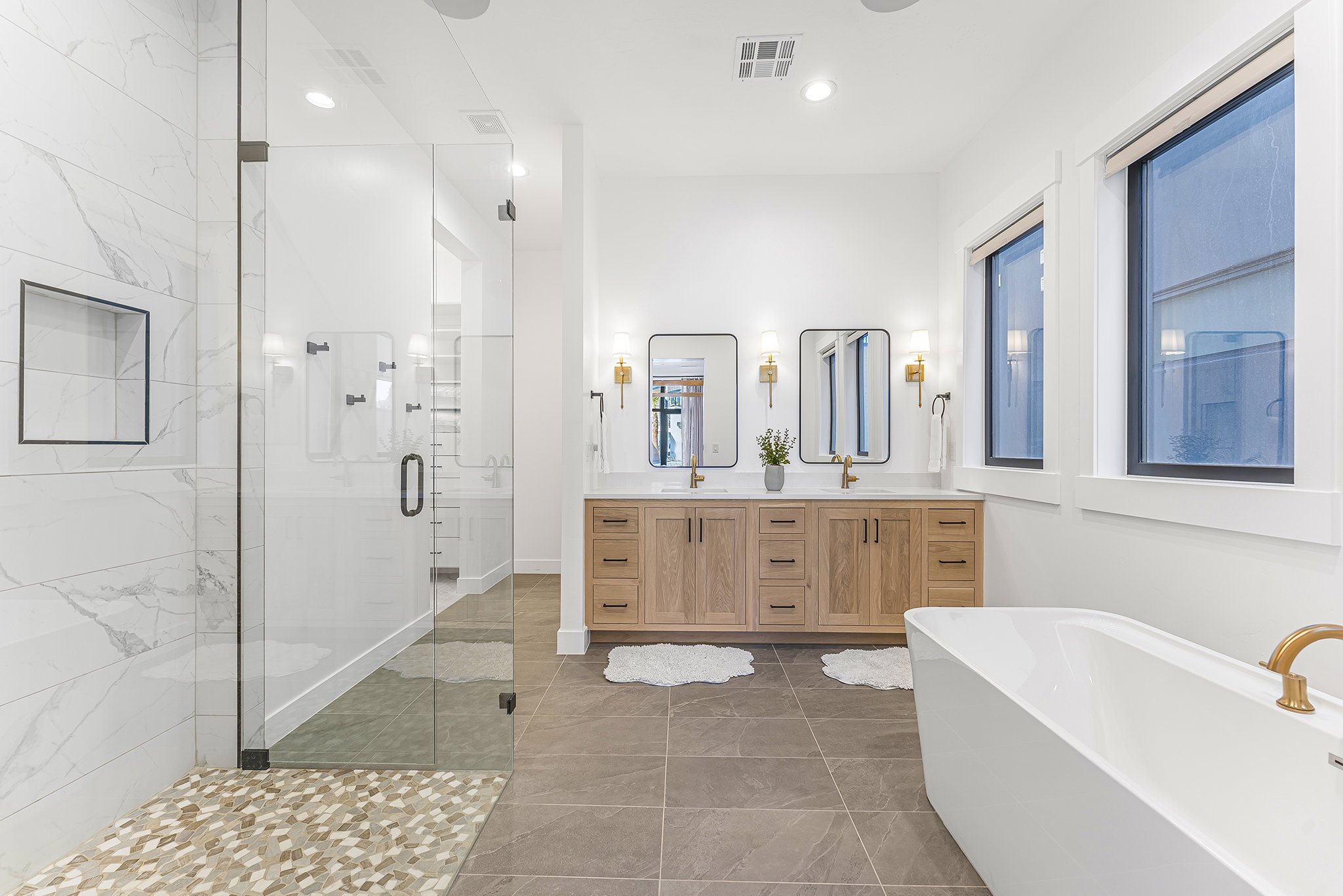 Spacious bathroom featuring a freestanding soaking tub, a dual vanity with wood-finish cabinetry, a large glass-enclosed shower with mosaic tile flooring, and two framed mirrors with sconce lighting