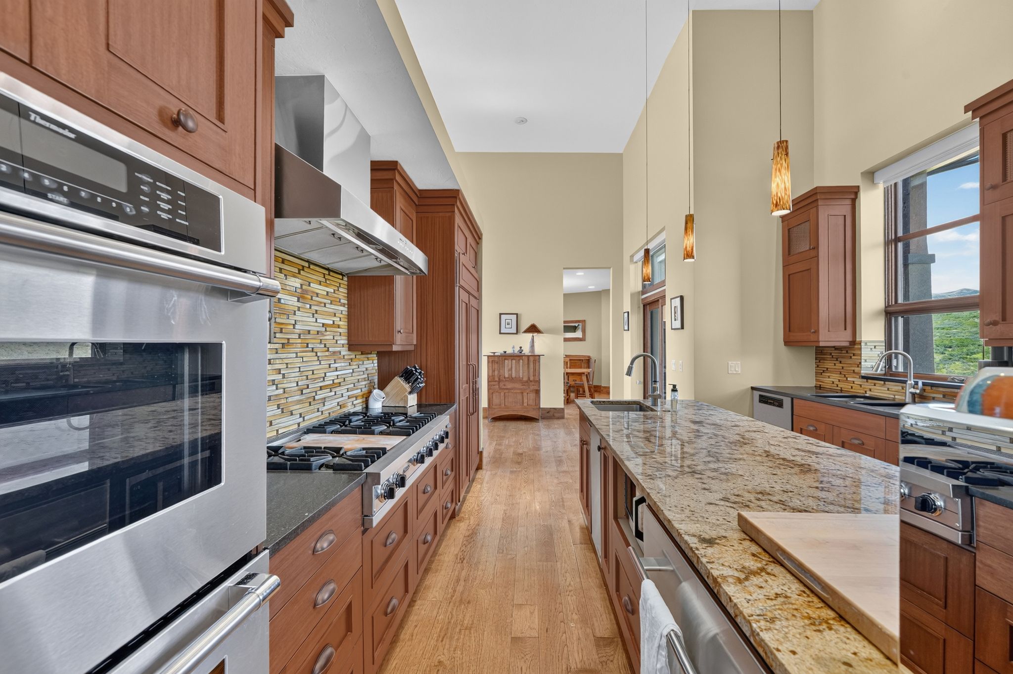 Gourmet kitchen featuring a large central island with an undermount sink, ample wood cabinetry, integrated stainless steel appliances, a tiled backsplash, and high ceilings with pendant lighting